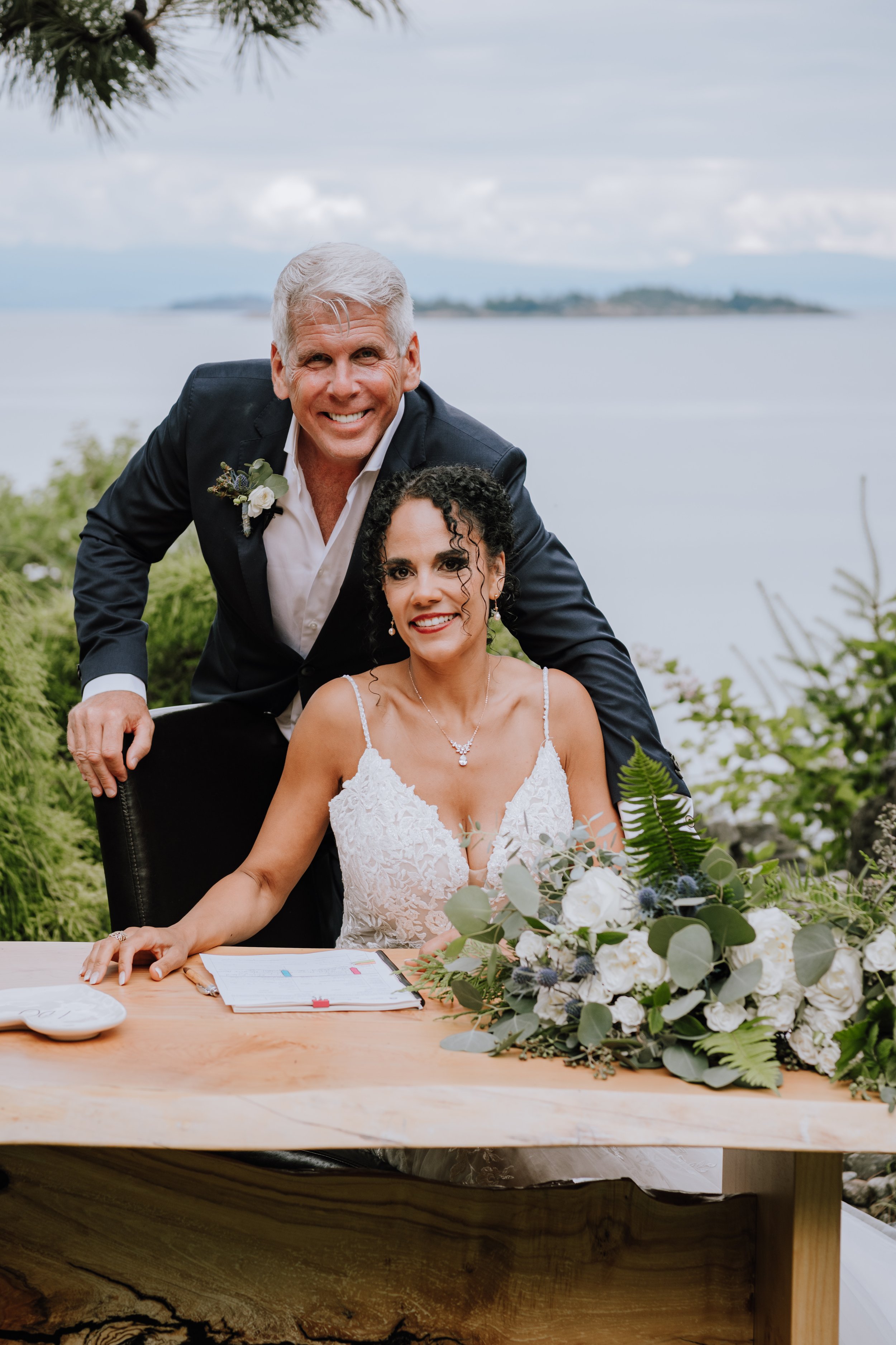 A bride and an older man, likely a groom or officiant, posing at an outdoor wedding by the water with a large bouquet of white flowers and greenery on a wooden table.
