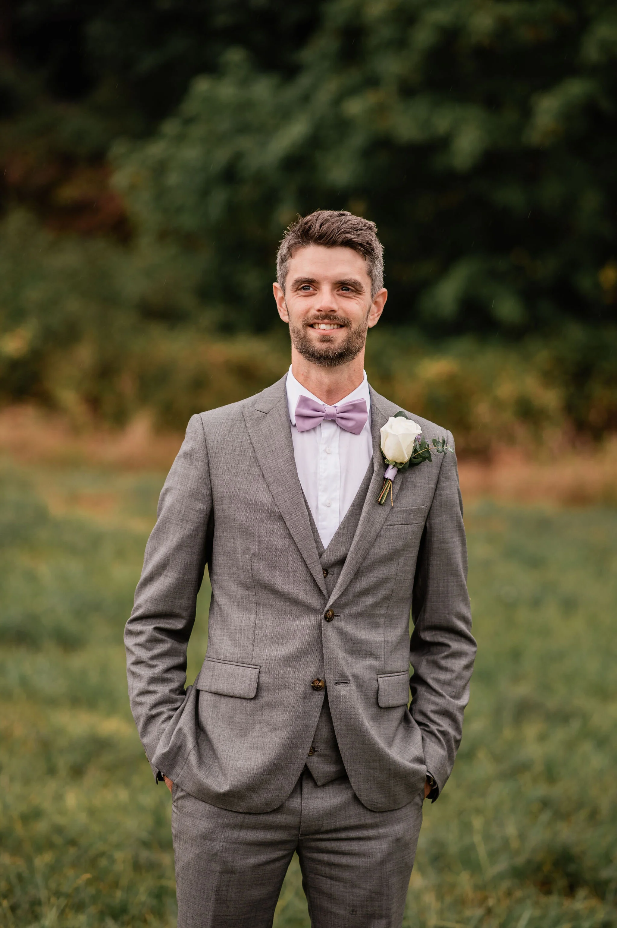 A man in a gray suit with a lavender bow tie and a white shirt, standing outdoors with trees in the background, smiling and with hands in his pockets.
