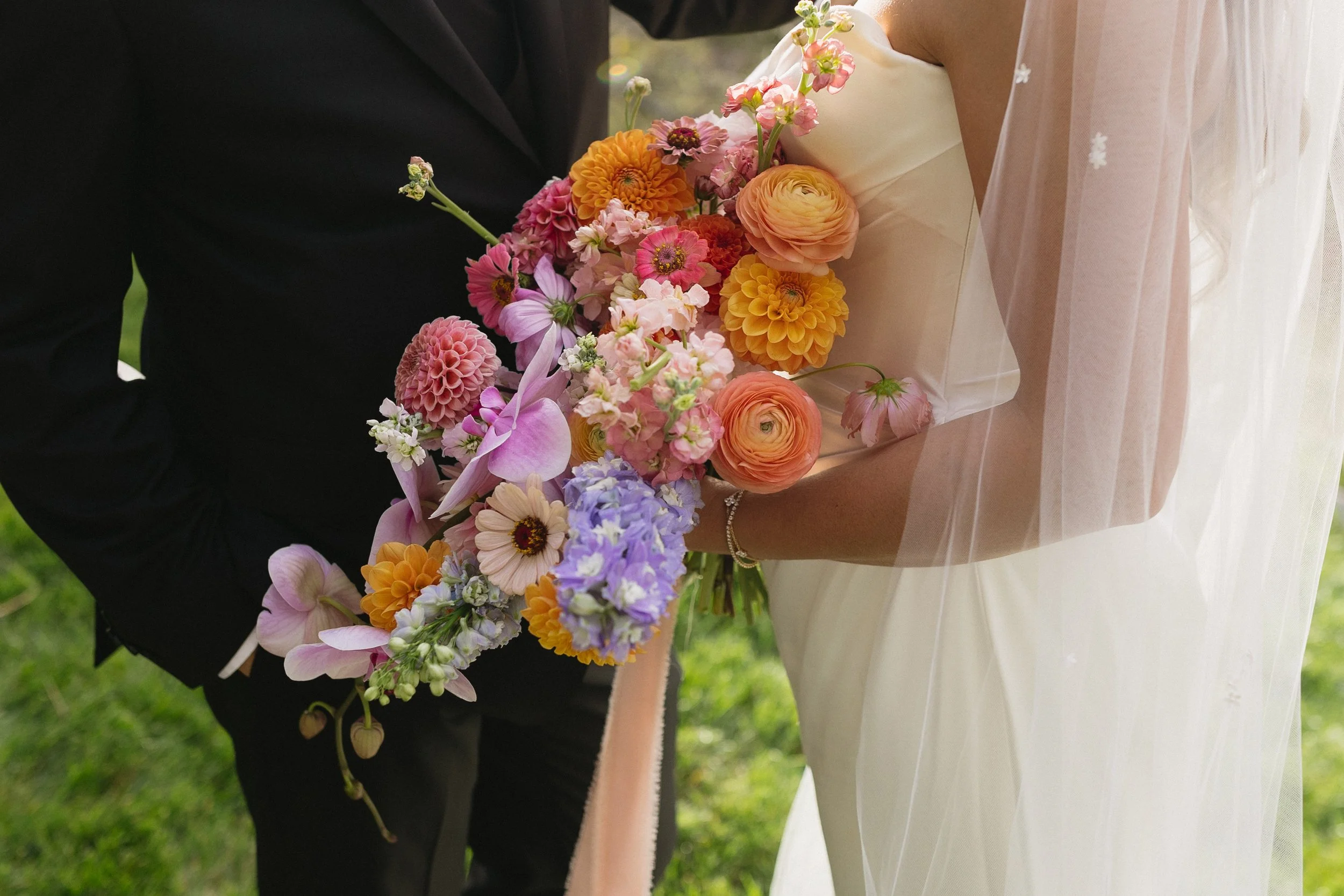 Close-up of a bride and groom holding a colorful bouquet of flowers, with the bride wearing a wedding dress and veil and the groom in a black suit, set outdoors on green grass.