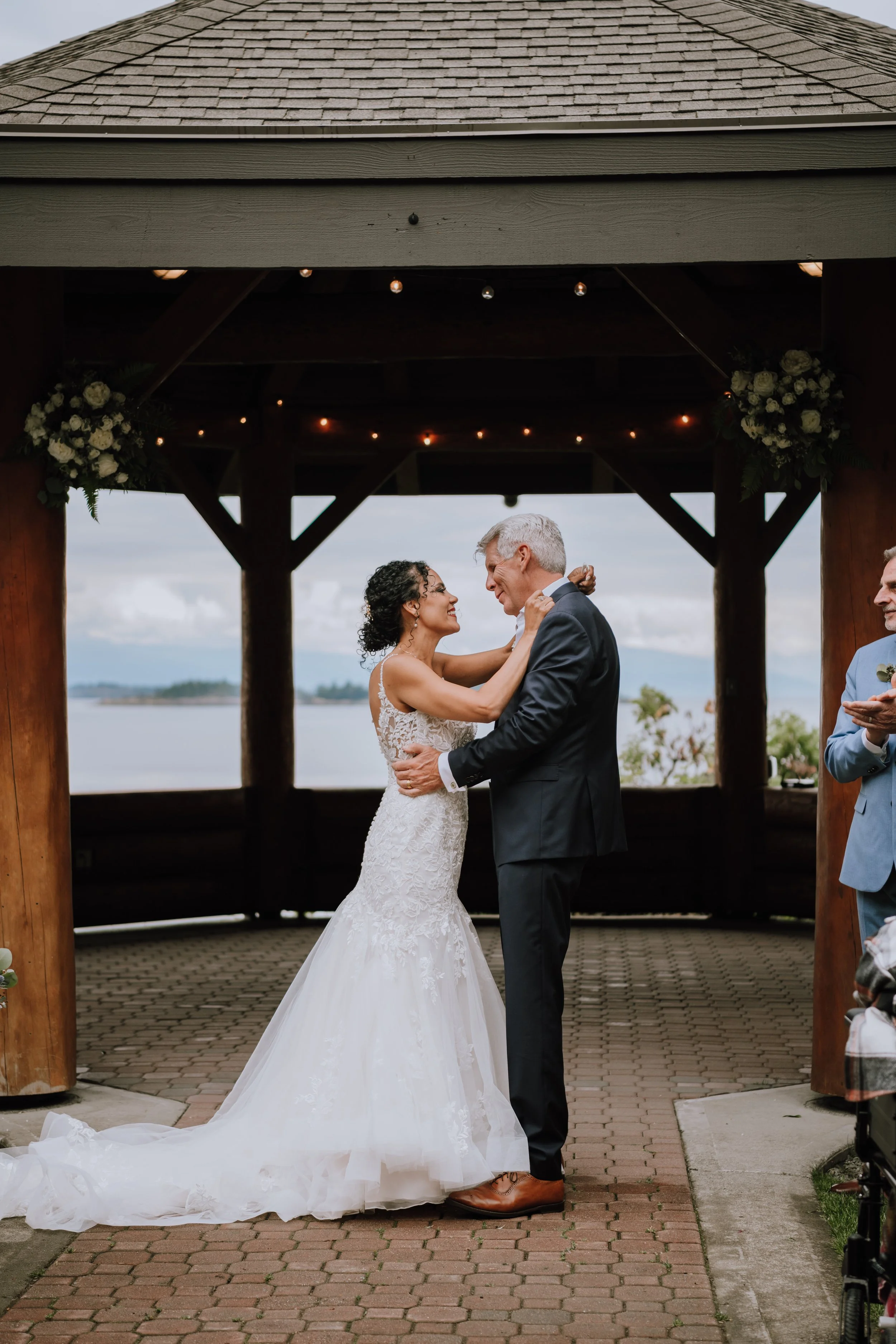 Couple having a wedding ceremony outdoors, with the bride in a white gown and the groom in a dark suit, dancing under a wooden pavilion with ocean in the background.