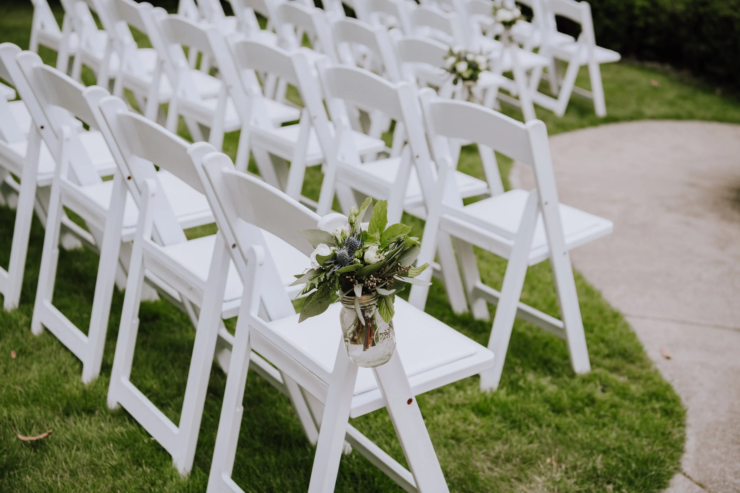 White folding chairs arranged outdoors for a wedding ceremony, decorated with a small bouquet of greenery and white flowers attached to one chair.