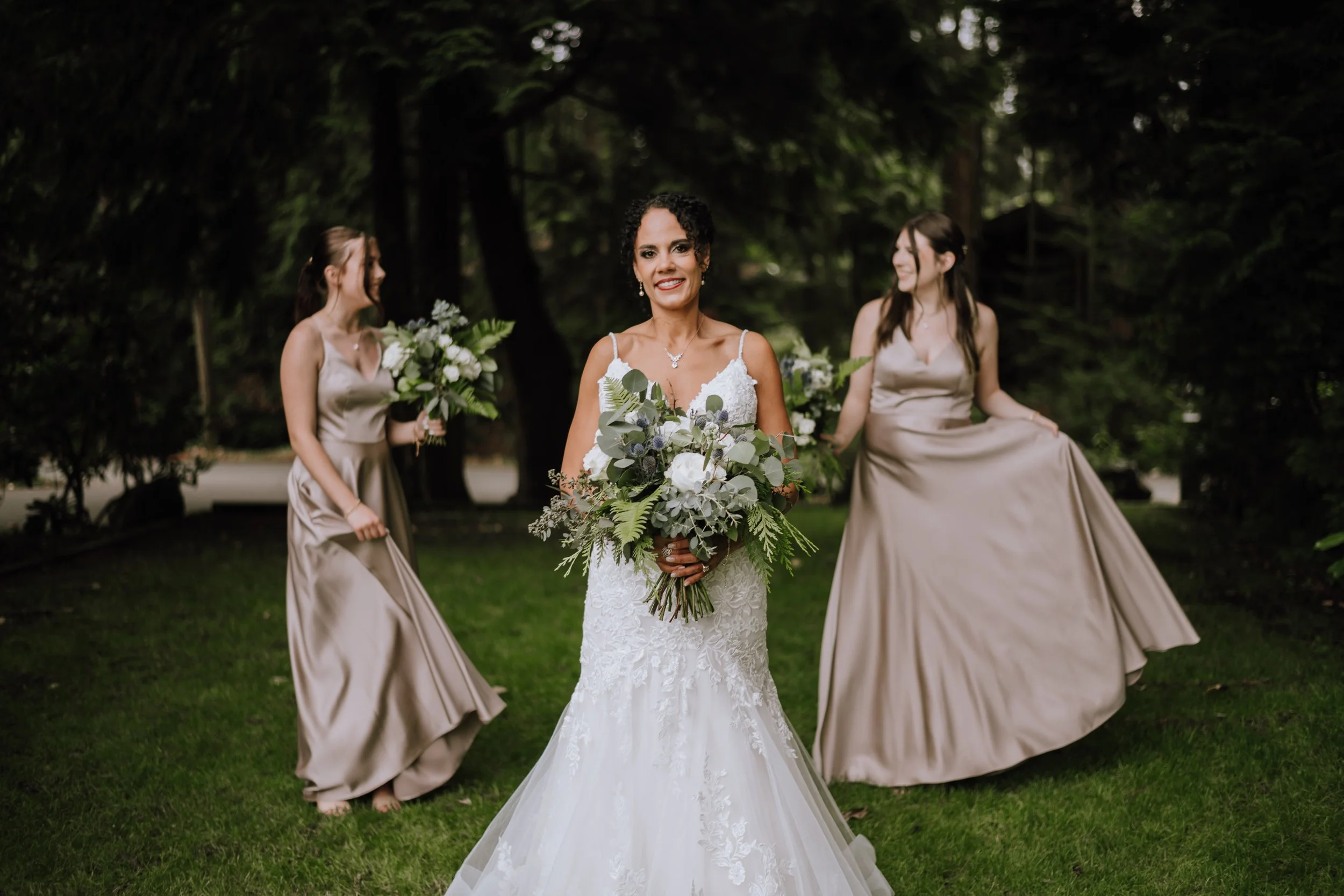 Bride in a white wedding dress holding a bouquet, flanked by two bridesmaids in taupe dresses holding bouquets, in a lush green outdoor setting.