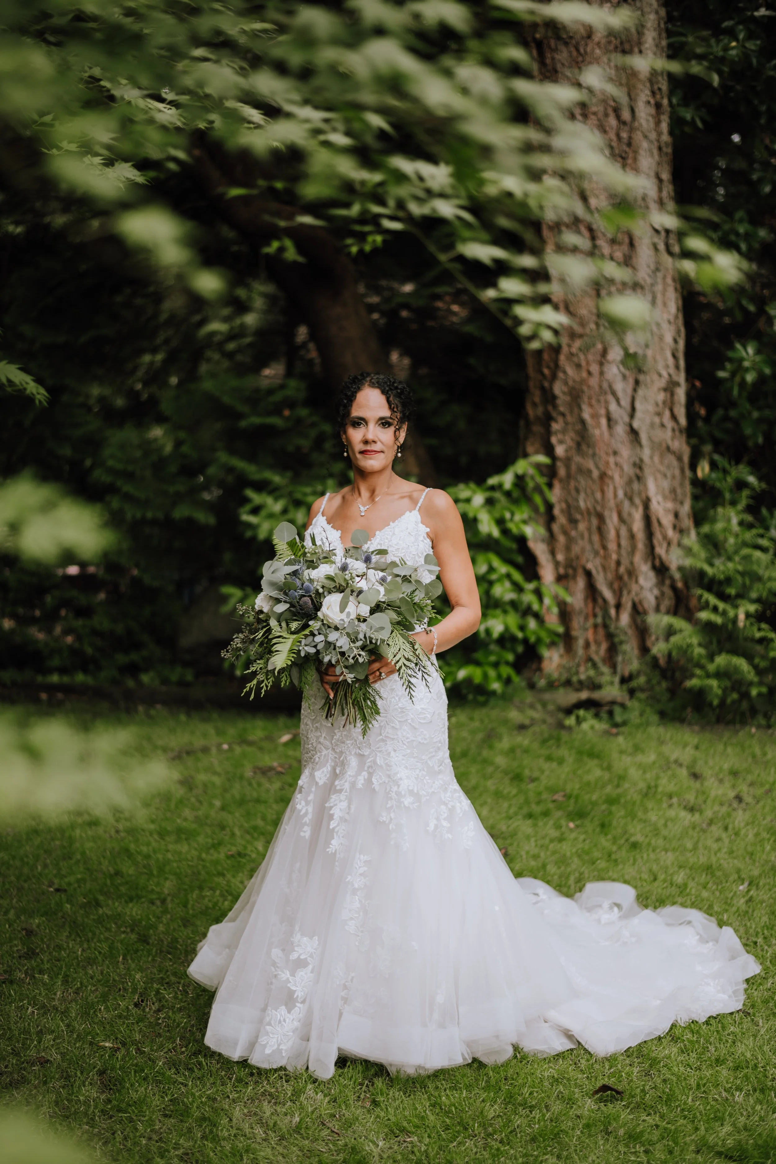 A bride in a white lace wedding gown holding a large bouquet of greenery and white flowers, standing on a grassy area in a forest with trees and greenery in the background.