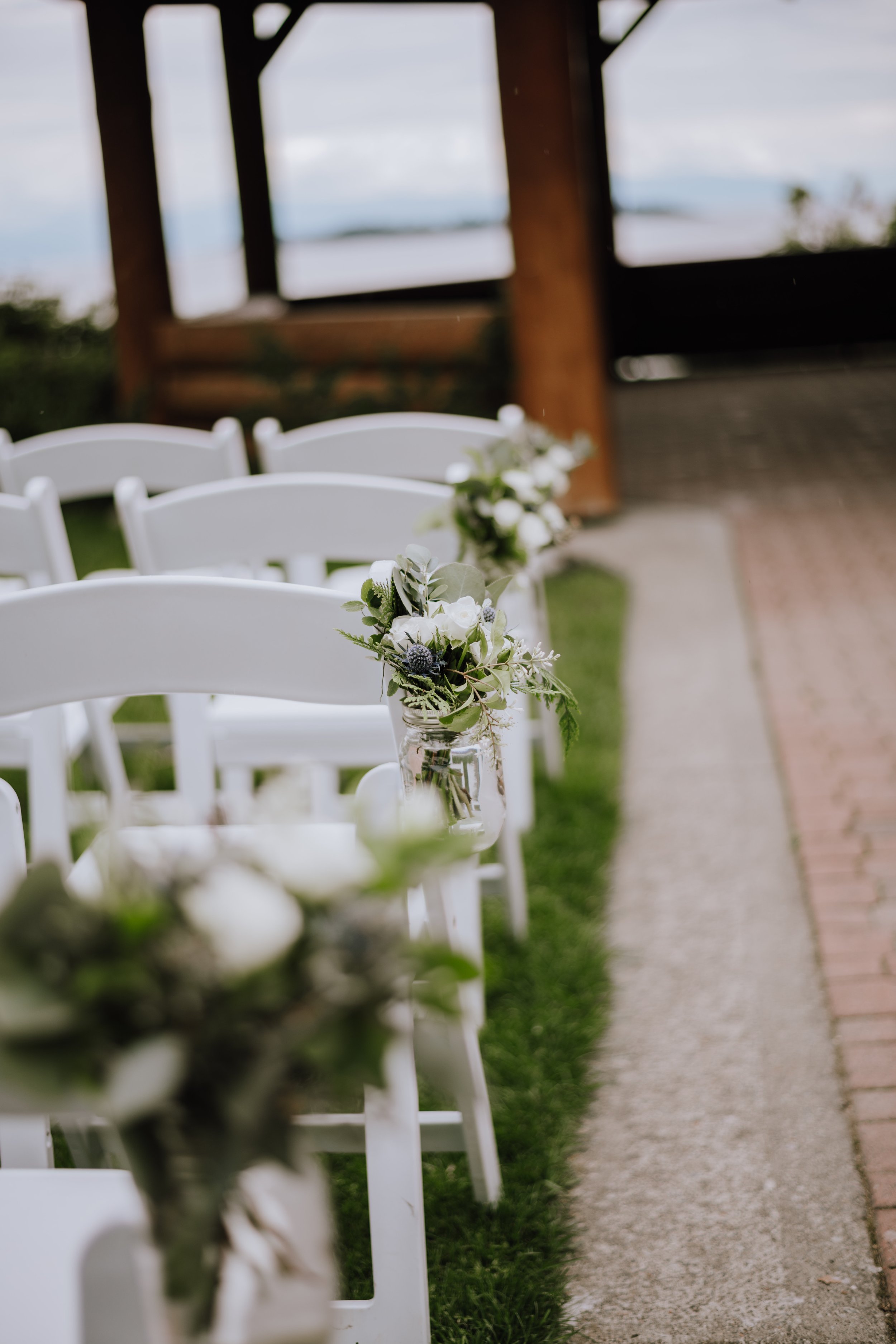 Wedding aisle decorated with white flowers in glass vases, white folding chairs on a grassy area, outdoor venue with wooden structure in background.