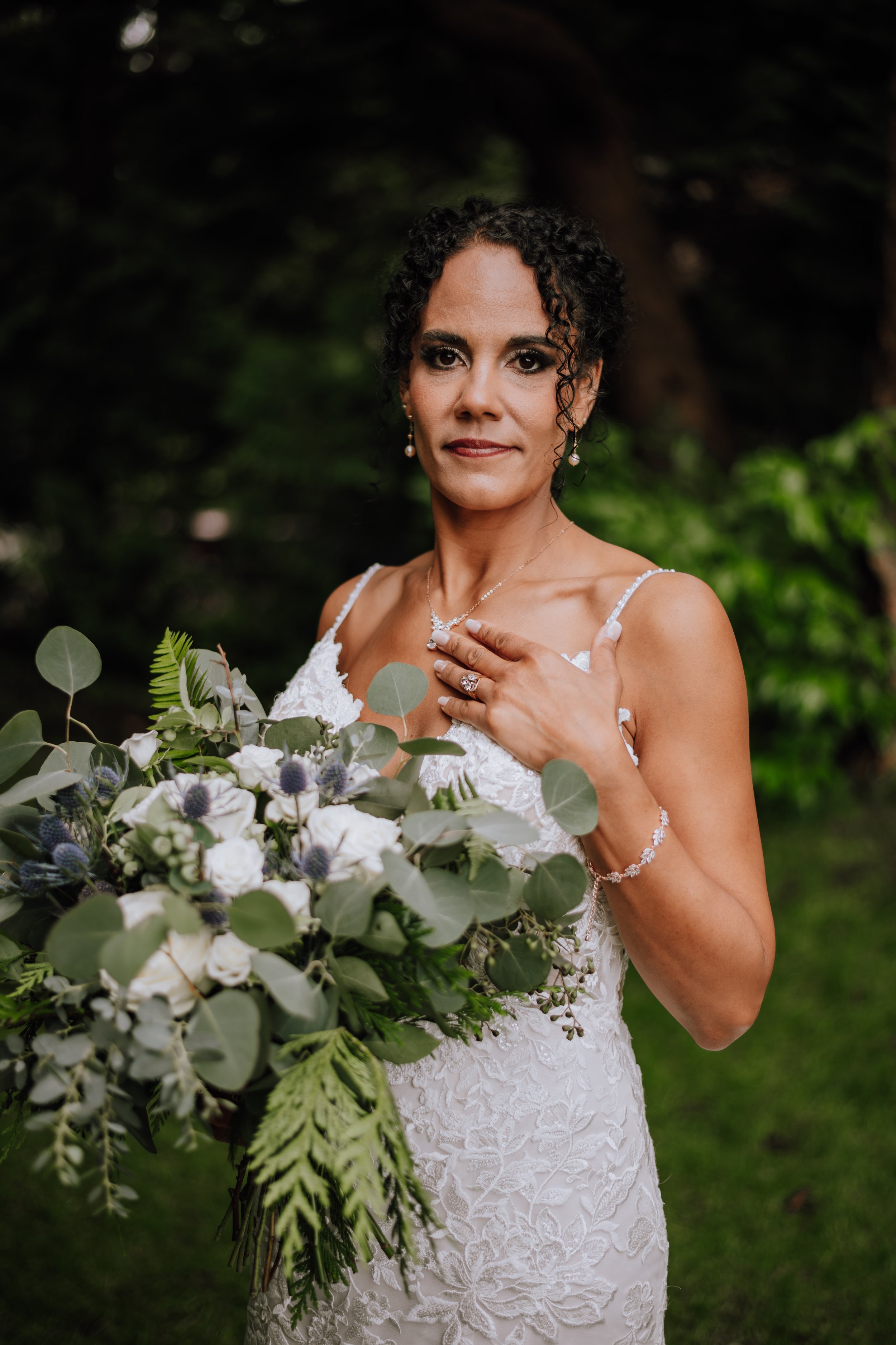 A bride in a white lace wedding dress holding a large bouquet of white and purple flowers and greenery, standing outdoors with trees in the background.