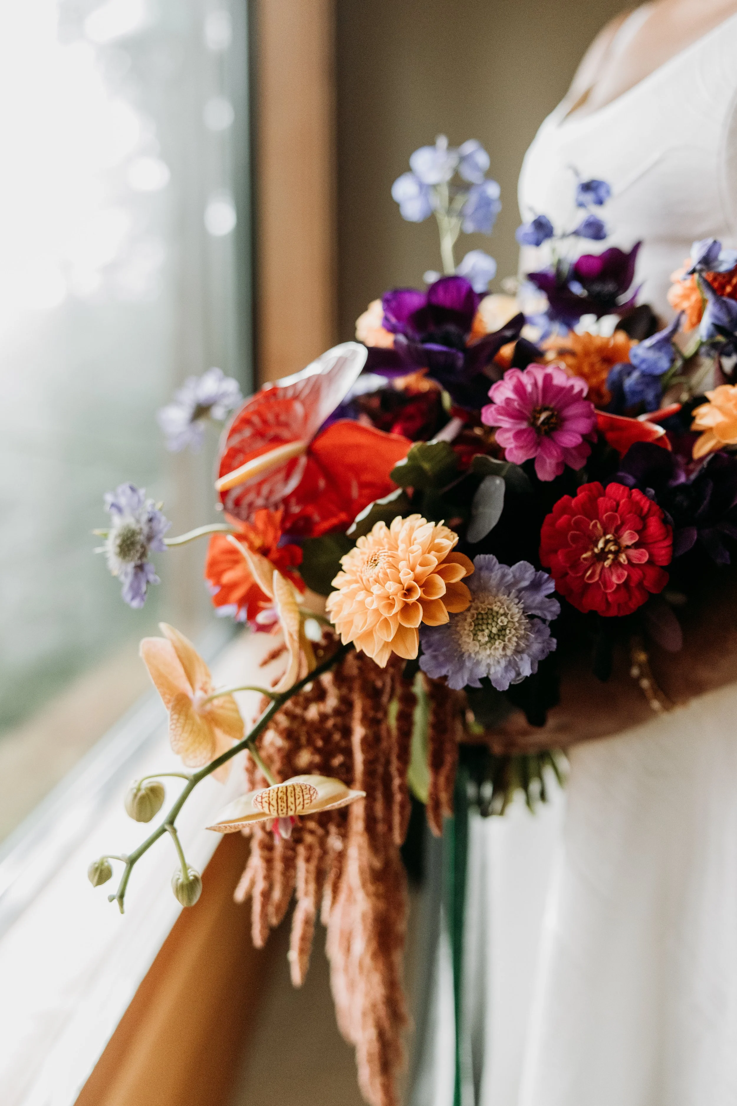 Close-up of a colorful bouquet of flowers with purple, pink, orange, and white blossoms, placed on a windowsill with a brown window frame in the background.