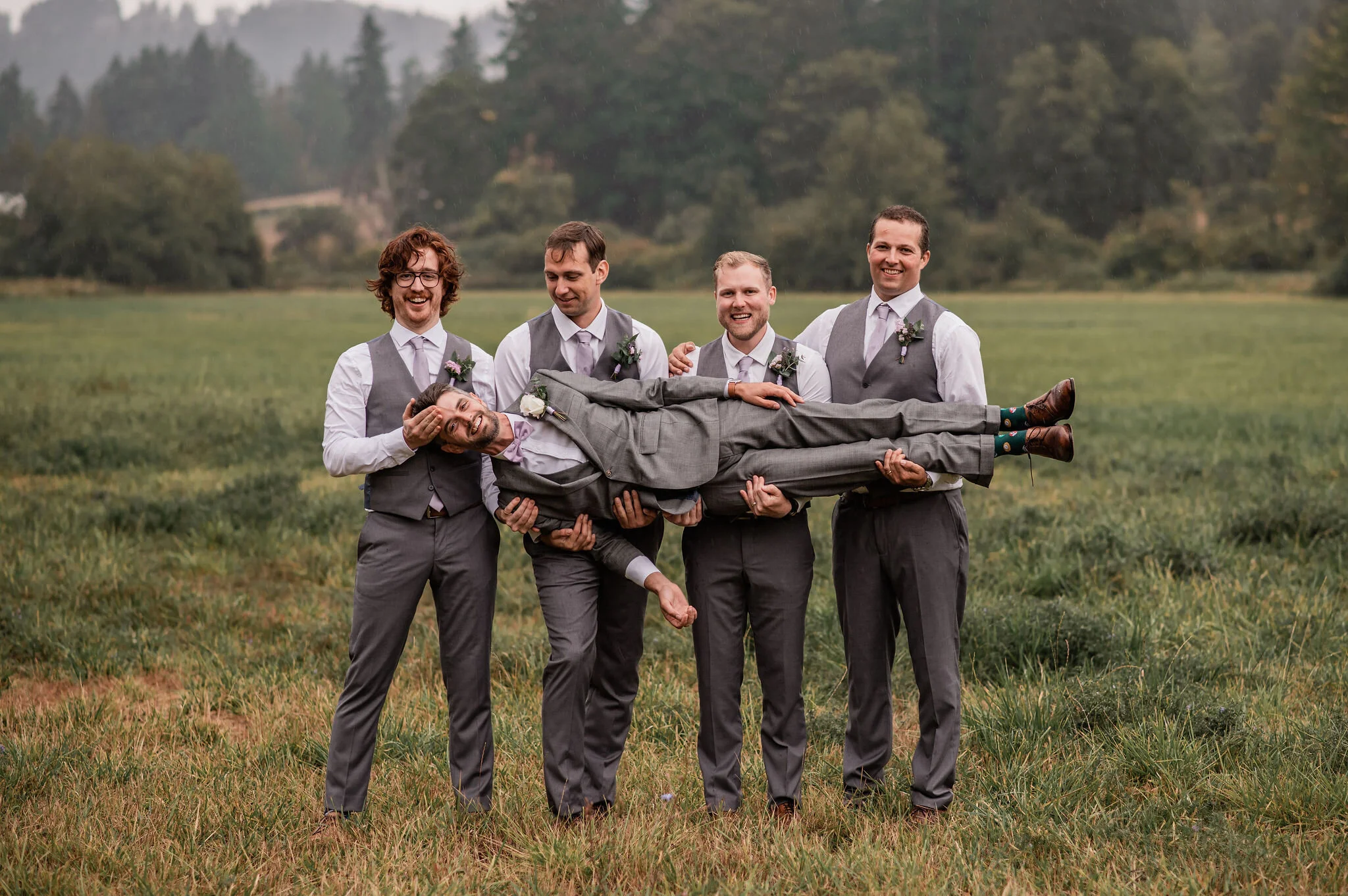 Groom and groomsmen holding a groom-lie down pose outdoors on a grassy field with trees and hills in the background.