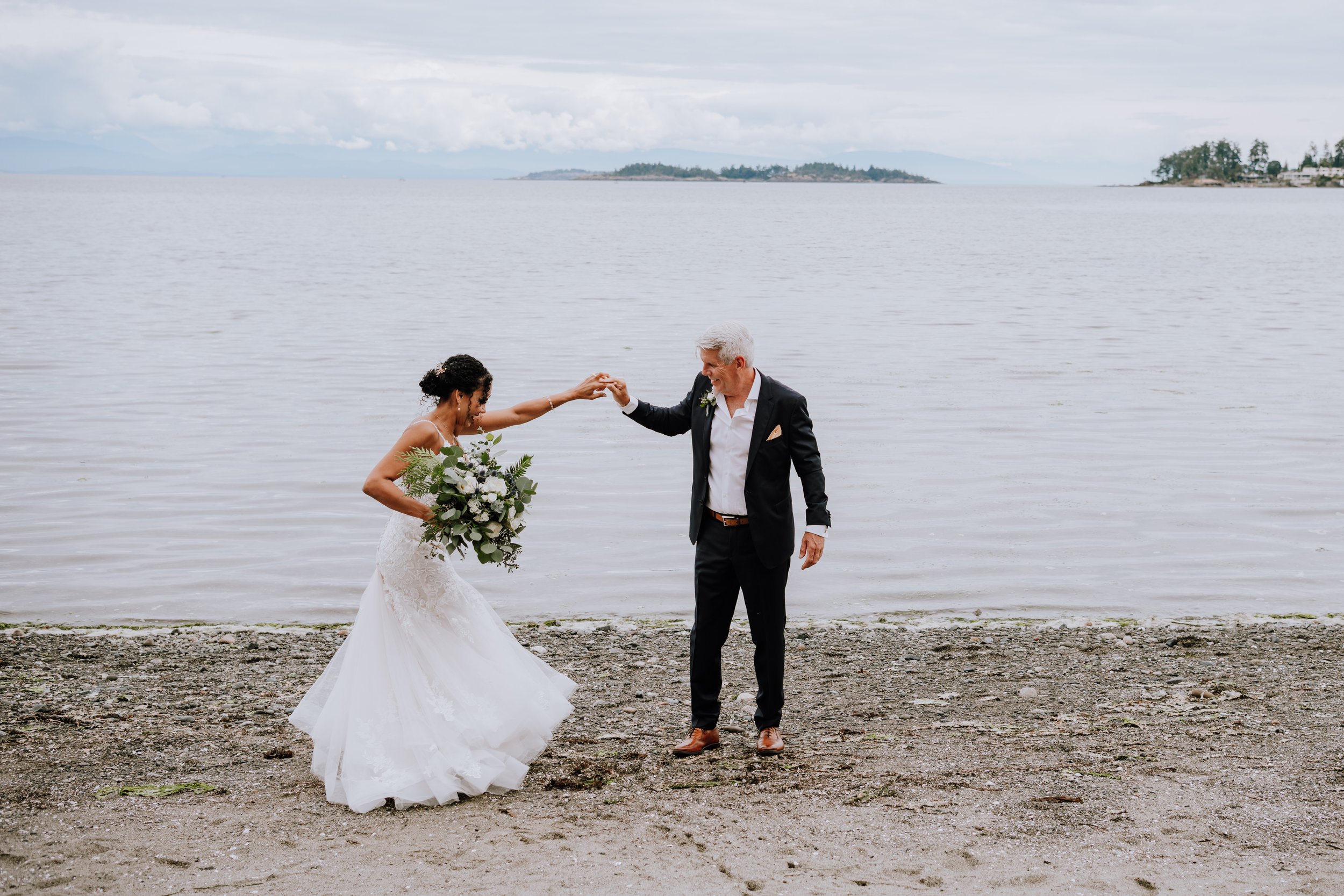 A bride and an older man, likely her father, dancing on a beach with water and islands in the background. The bride is wearing a white wedding gown and holding a bouquet, and the man is dressed in a black suit with a boutonniere.