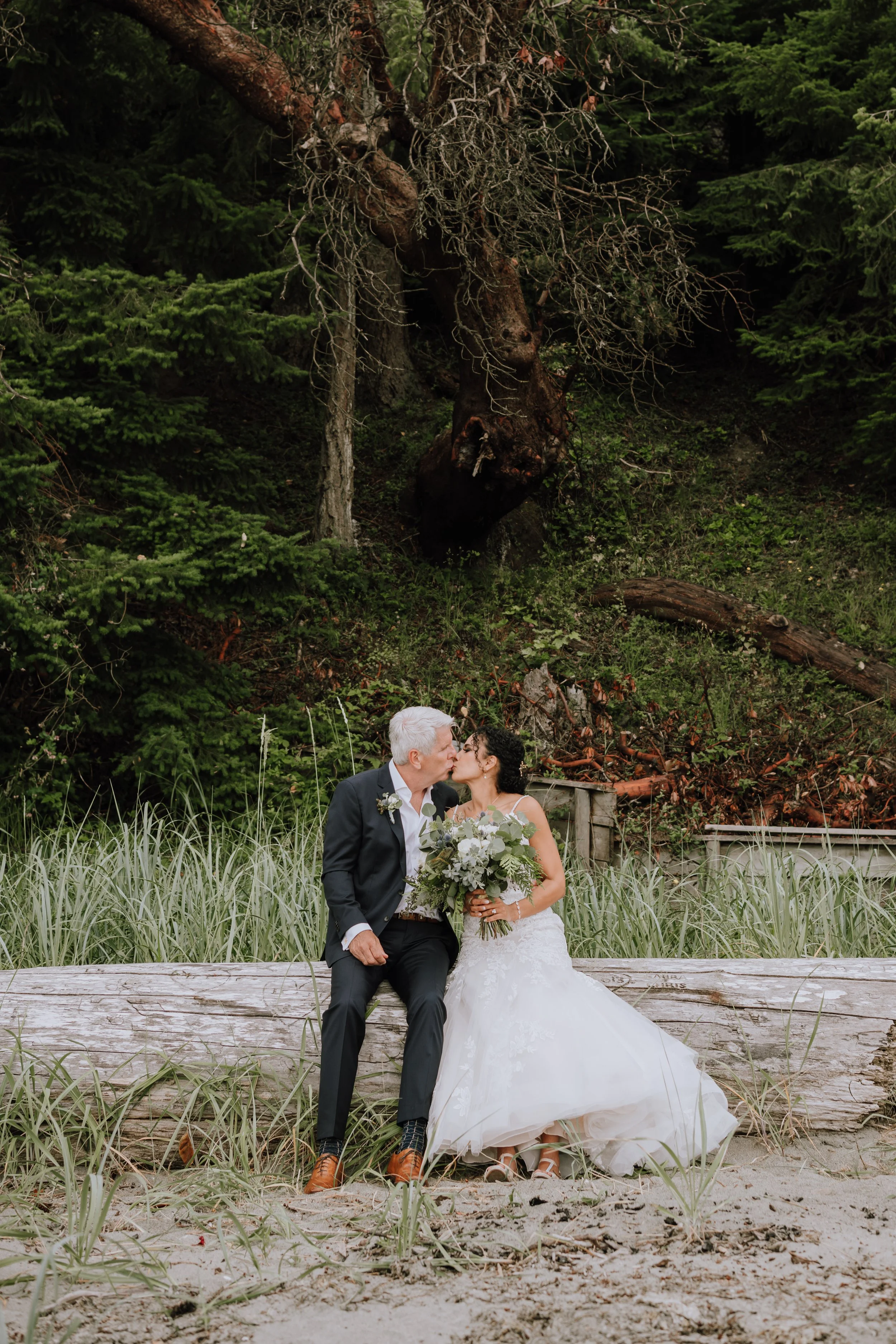 A bride and groom sit on a log on a sandy beach, sharing a kiss, with a large fallen tree and dense green forest in the background.