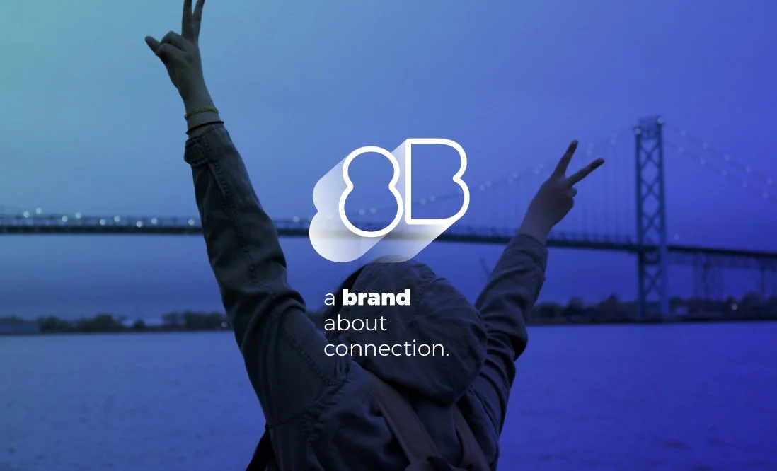 A person with arms raised making peace signs against a blue and purple sunset over a body of water, with a bridge in the distance. Overlay text reads "a brand about connection."