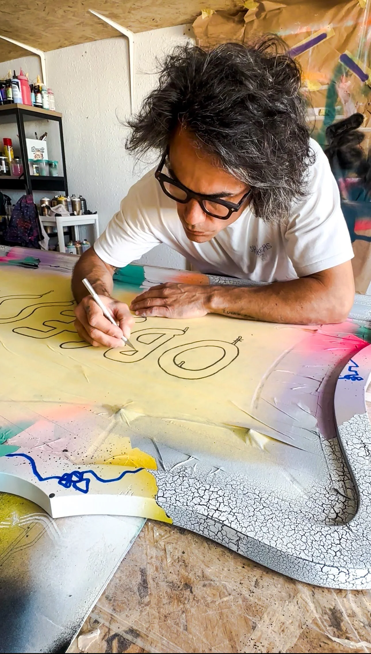 An artist with gray hair and glasses is drawing on a colorful, abstract painting on a table. The background shows art supplies on shelves, a backpack, and protective paper taped to the walls.