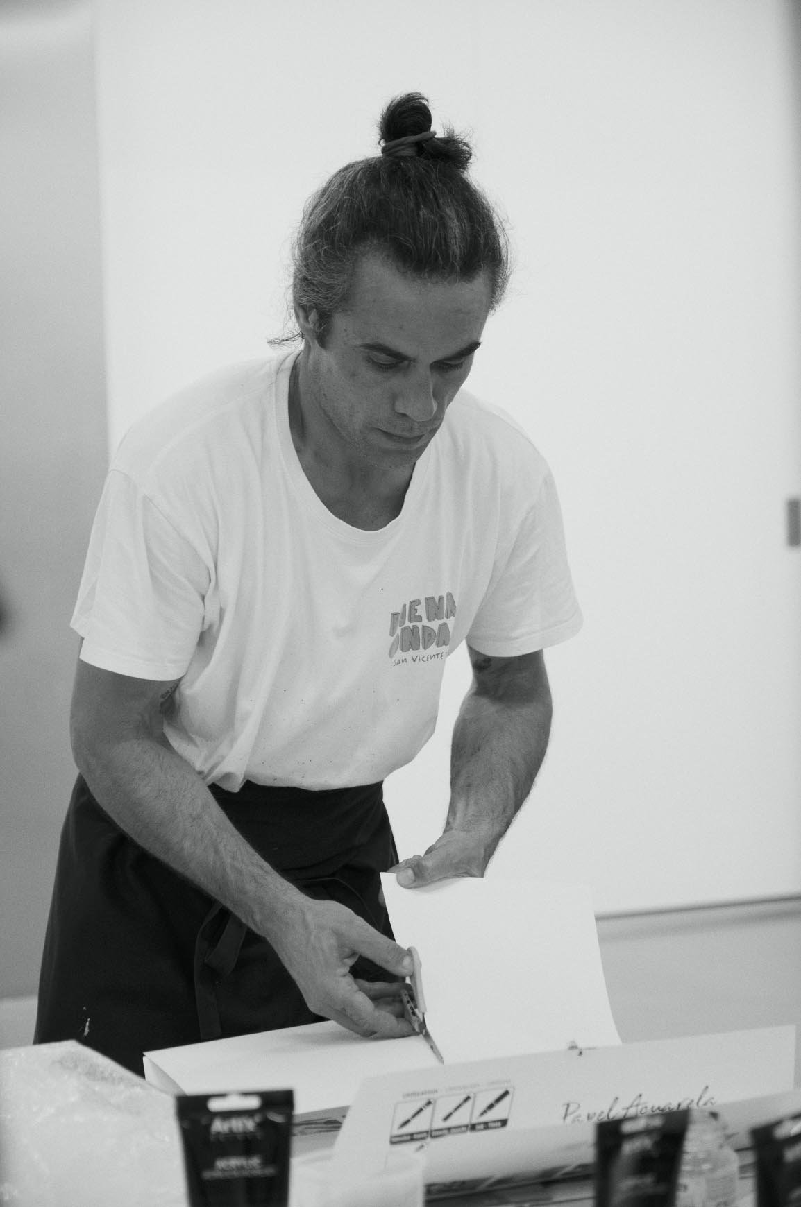 A man with a man bun hairstyle wearing a white t-shirt, standing at a table, holding and possibly folding a piece of paper or cardboard, in an indoor setting with plain walls.