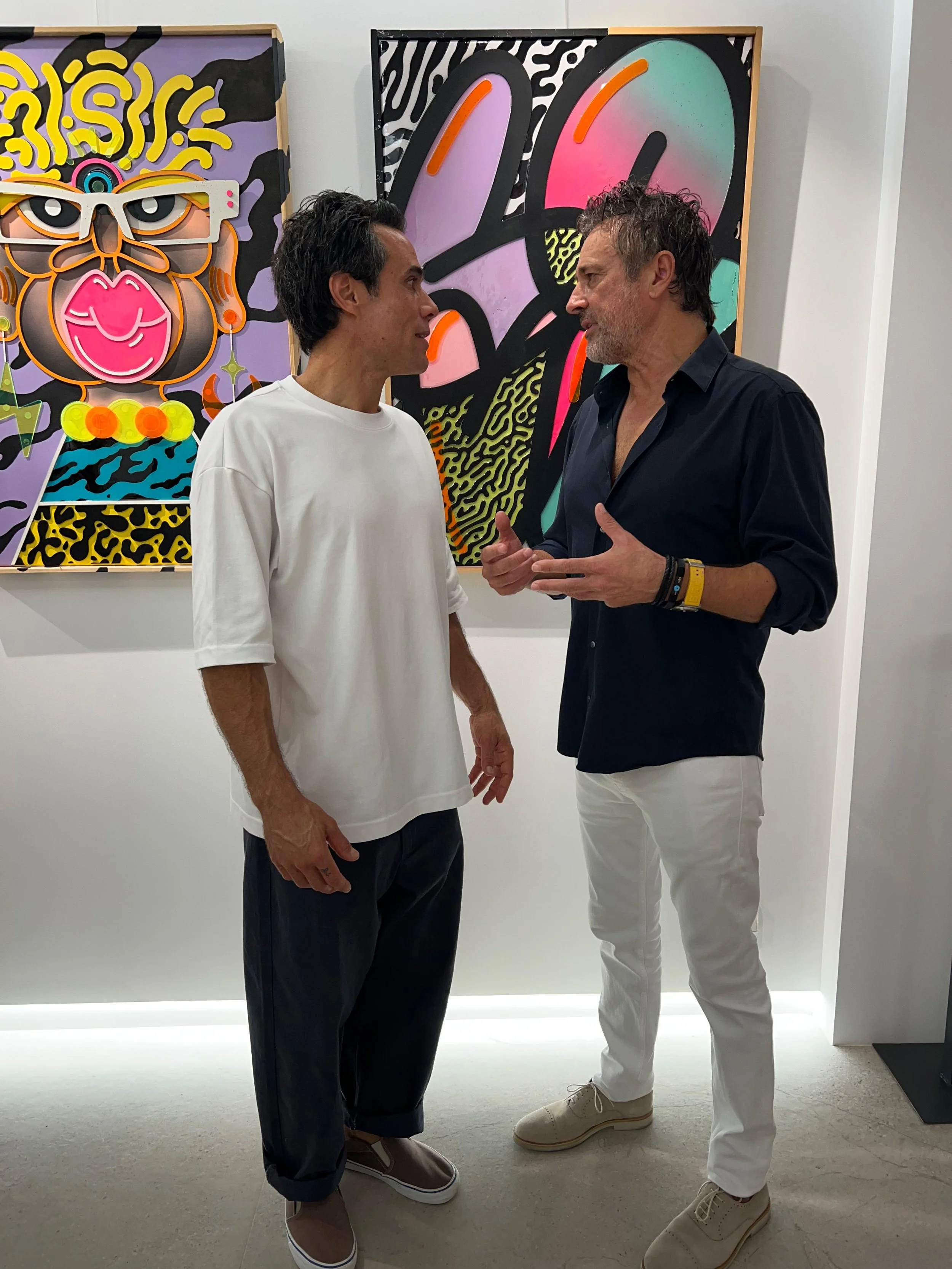 Two men are talking while standing in front of two colorful artworks at an art gallery. The man on the left is wearing a white T-shirt and dark pants, and the man on the right is wearing a dark shirt and white pants.