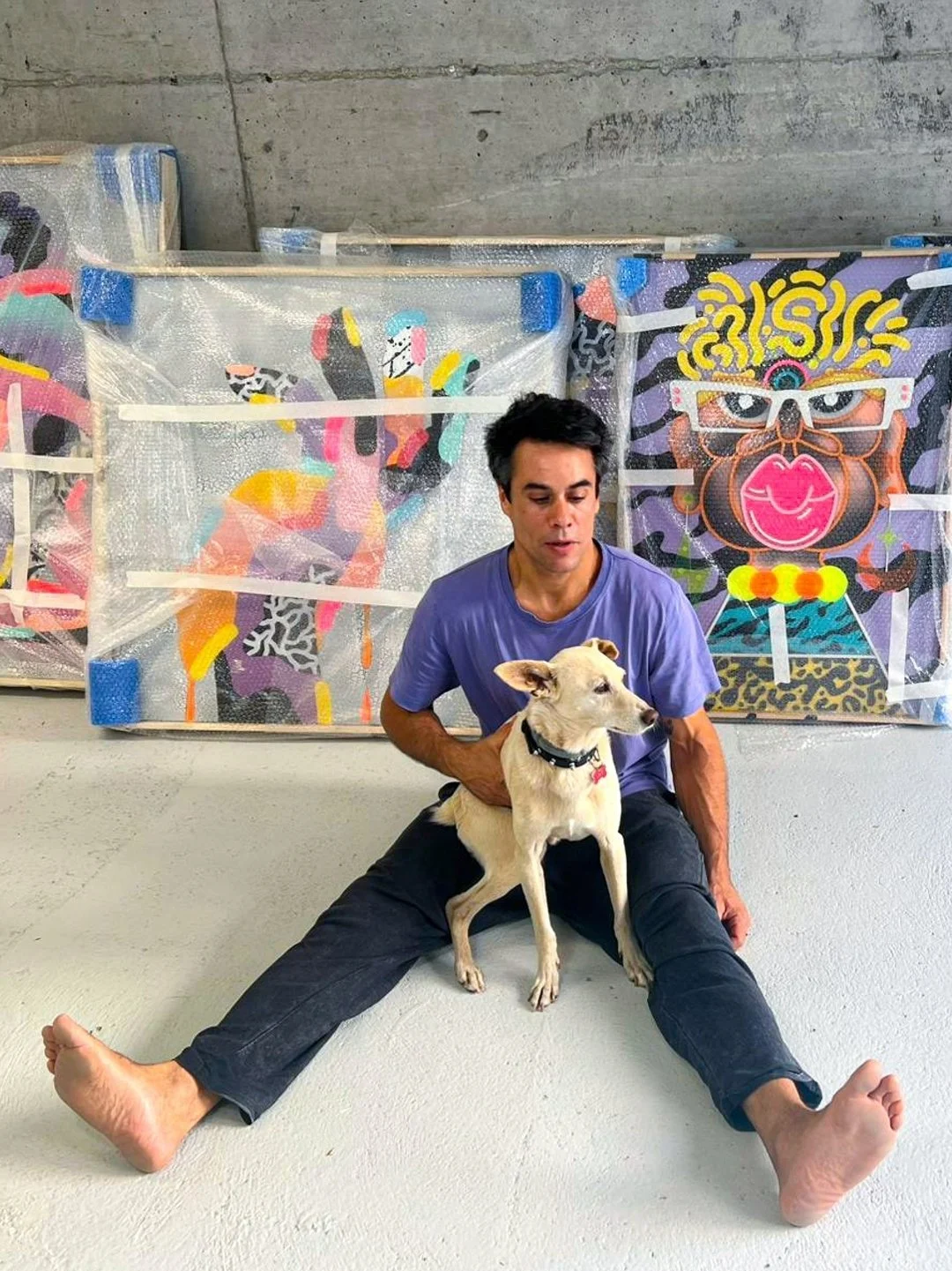 A man sitting on the floor with a dog sitting on his lap, in front of colorful abstract paintings. The man has dark hair, and is wearing a purple T-shirt and black pants. The background features concrete walls and floor.