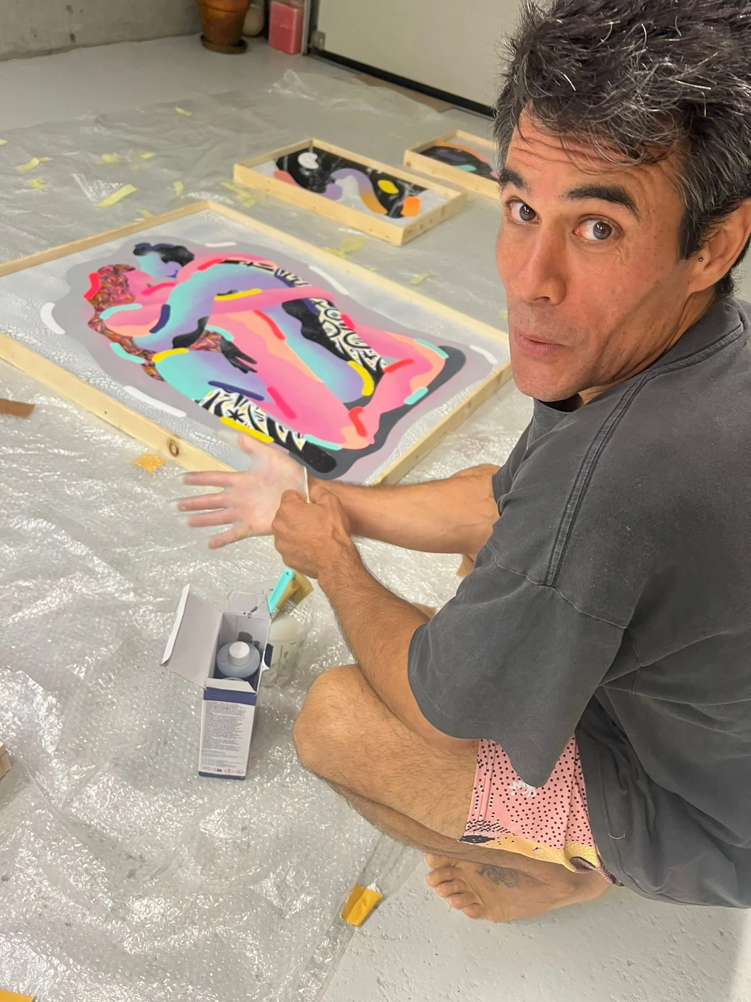 A man is sitting on the floor in front of a colorful abstract painting. He is holding the painting and looking at the camera. There are art supplies and wooden frames on the floor, covered with a plastic sheet.