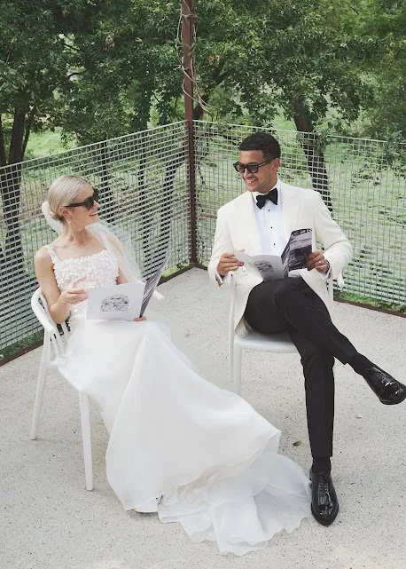 A bride and groom sitting outdoors on white chairs, dressed in wedding attire, smiling at each other, holding papers, with green trees and a metal fence in the background.