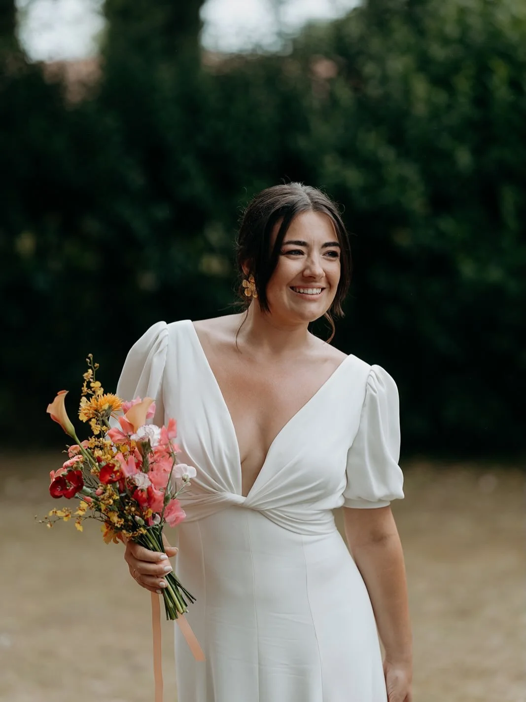 A woman in a white dress holding a colorful bouquet of flowers outdoors, smiling with dark hair and earrings.
