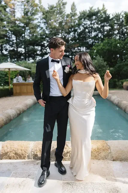 A bride and groom in wedding attire standing by a pool outdoors, smiling and looking at each other.