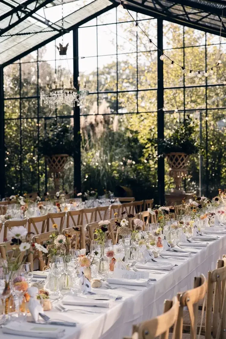 Long banquet table set with white tablecloth, floral centerpieces, wine glasses, and napkins inside a glass greenhouse with hanging string lights and chandeliers.