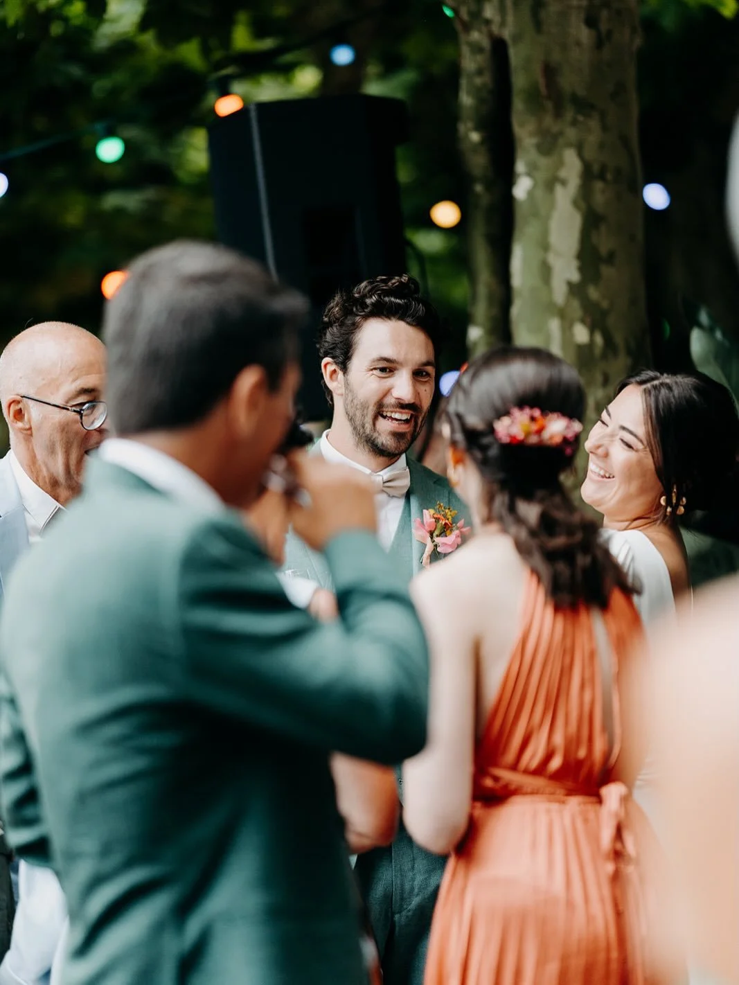 A group of people at an outdoor wedding reception, smiling and engaged in conversation, with a man in a suit at the center and a woman wearing a flower hair accessory.