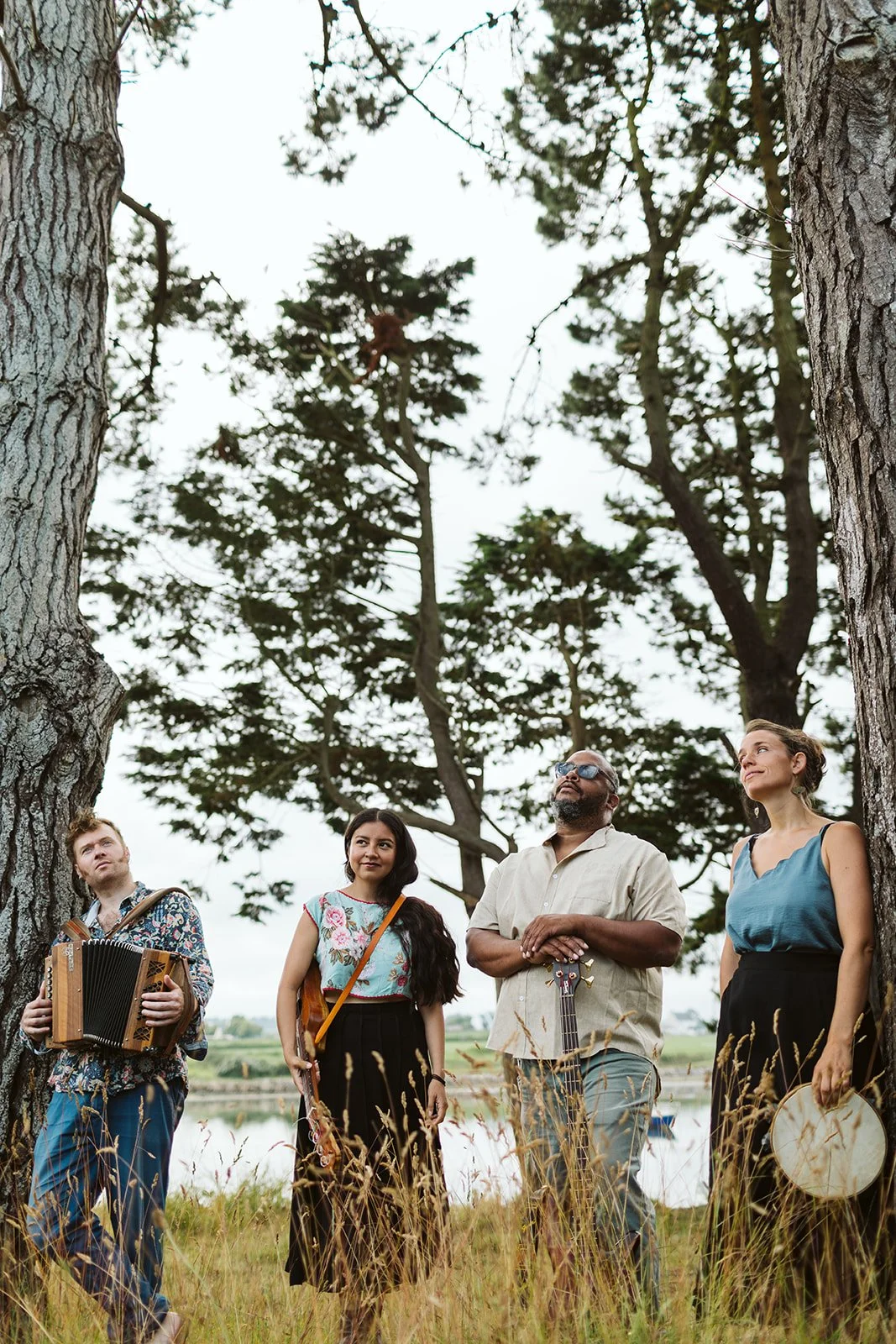 Quatre personnes jouent de la musique dans un champ près d'un lac, entourées d'arbres, un homme avec un accordéon, une femme avec une guitare, un autre homme avec une guitare et une femme avec un tambourin.