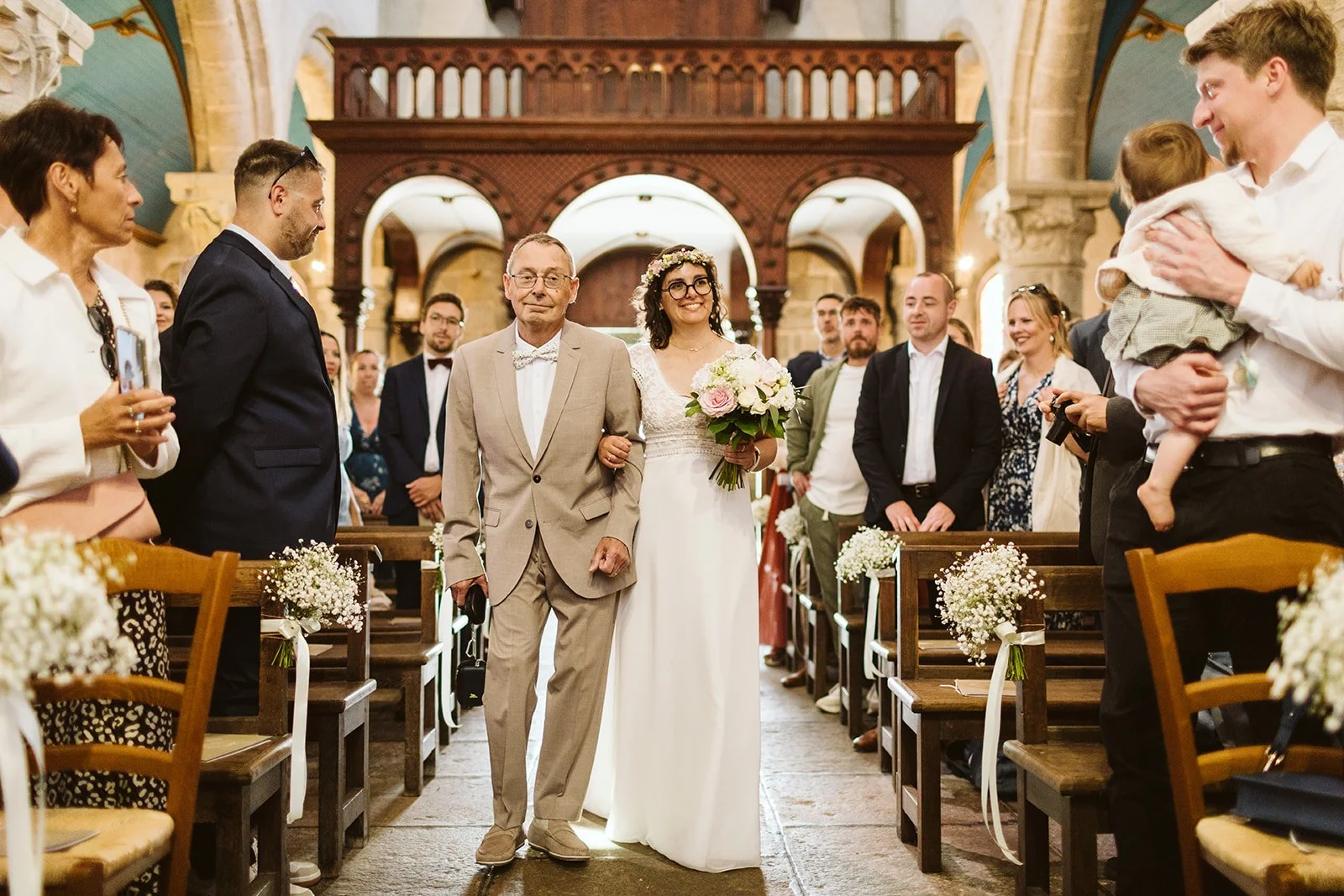 Une mariée souriante en robe blanche et couronne de fleurs de mariage, escortée par un homme âgé en costume beige, dans une église décorée, entourée de famille et amis lors d'une cérémonie nuptiale.