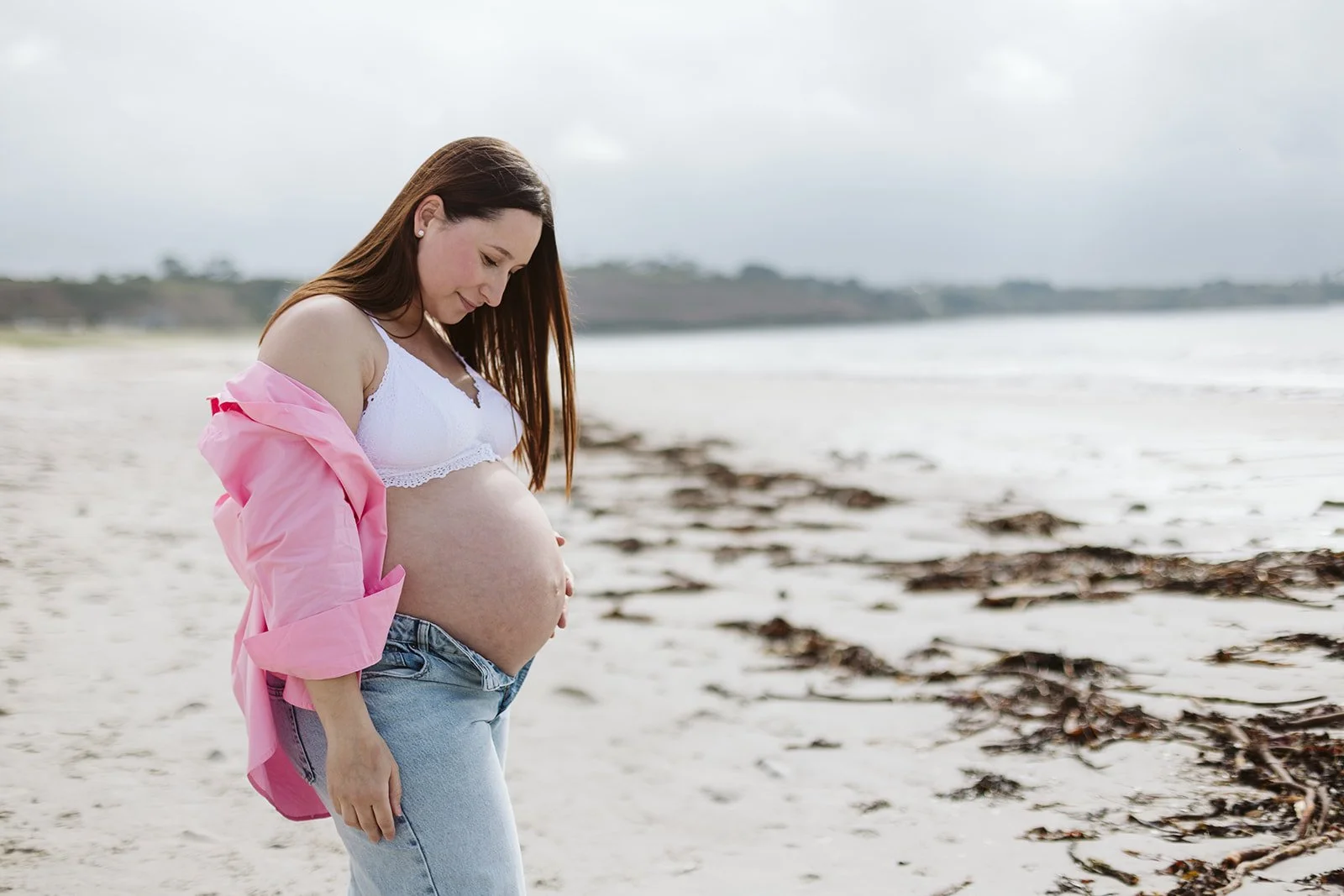 Jeune femme enceinte assise sur la plage, portant un haut blanc, une chemise rose défaite et un jean, regardant son ventre