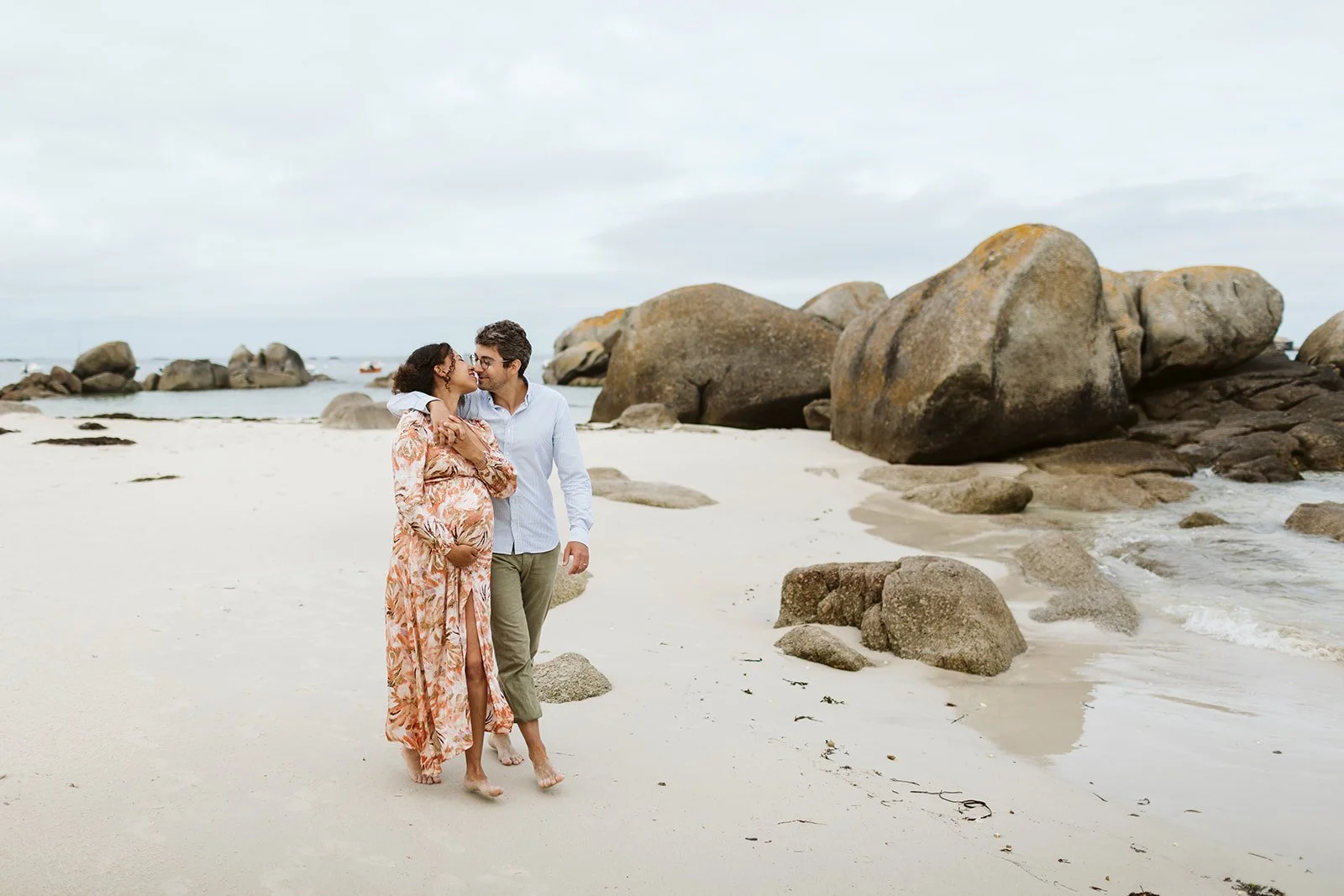 Un couple marche main dans la main sur la plage, l'homme caresse le ventre de la femme enceinte, avec des gros rochers en arrière-plan et un ciel nuageux.