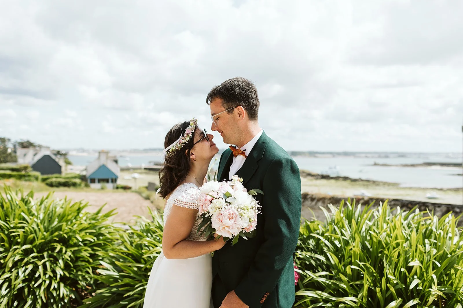 Un couple de mariés, une femme habillée en robe blanche et un homme en costume vert, se regardent avec amour en extérieur, la femme tenant un bouquet de fleurs pink et blanc.