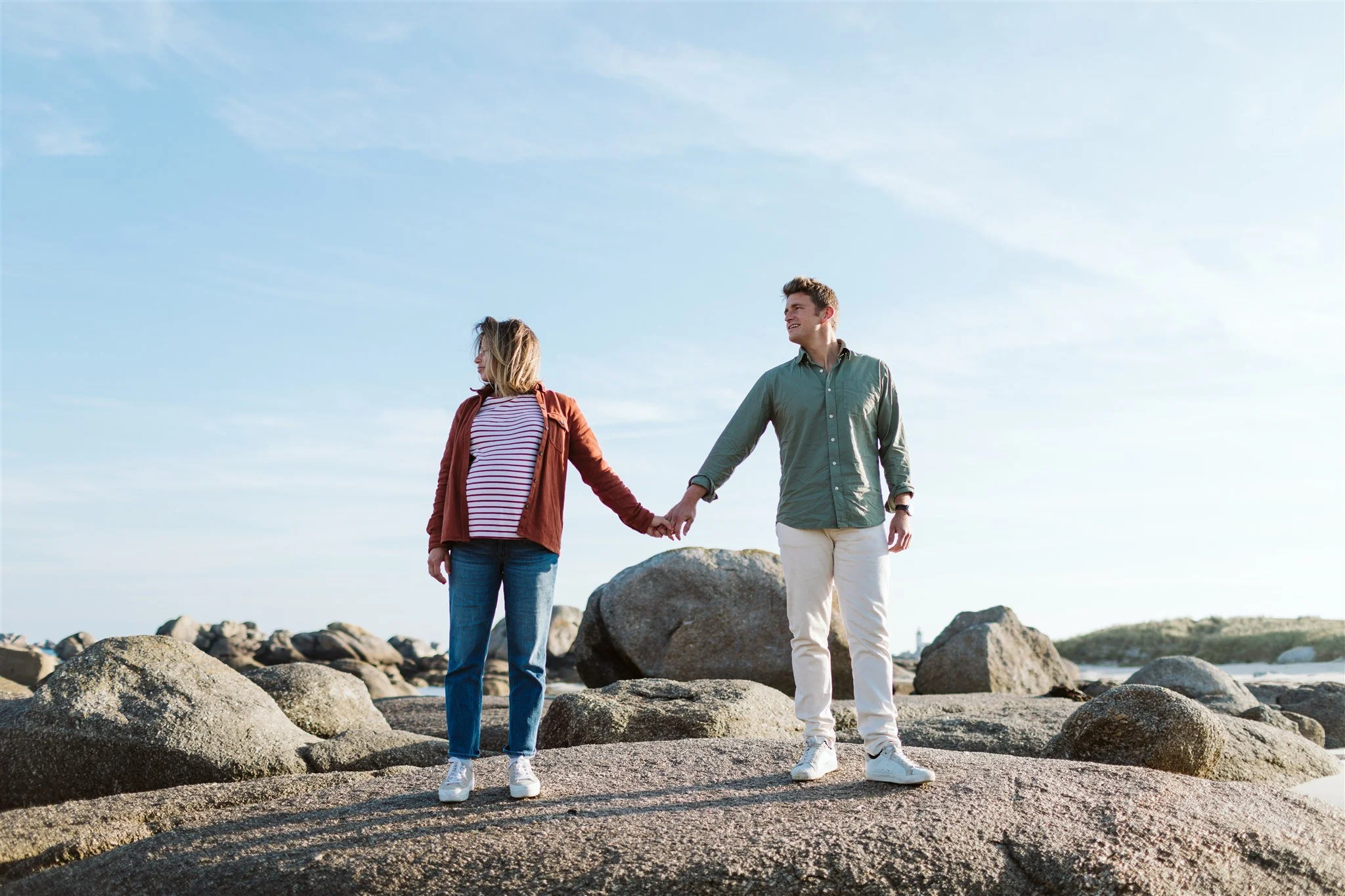 Un couple debout sur des rochers près de la mer, se tenant par la main, avec un ciel clair et des rochers en arrière-plan.