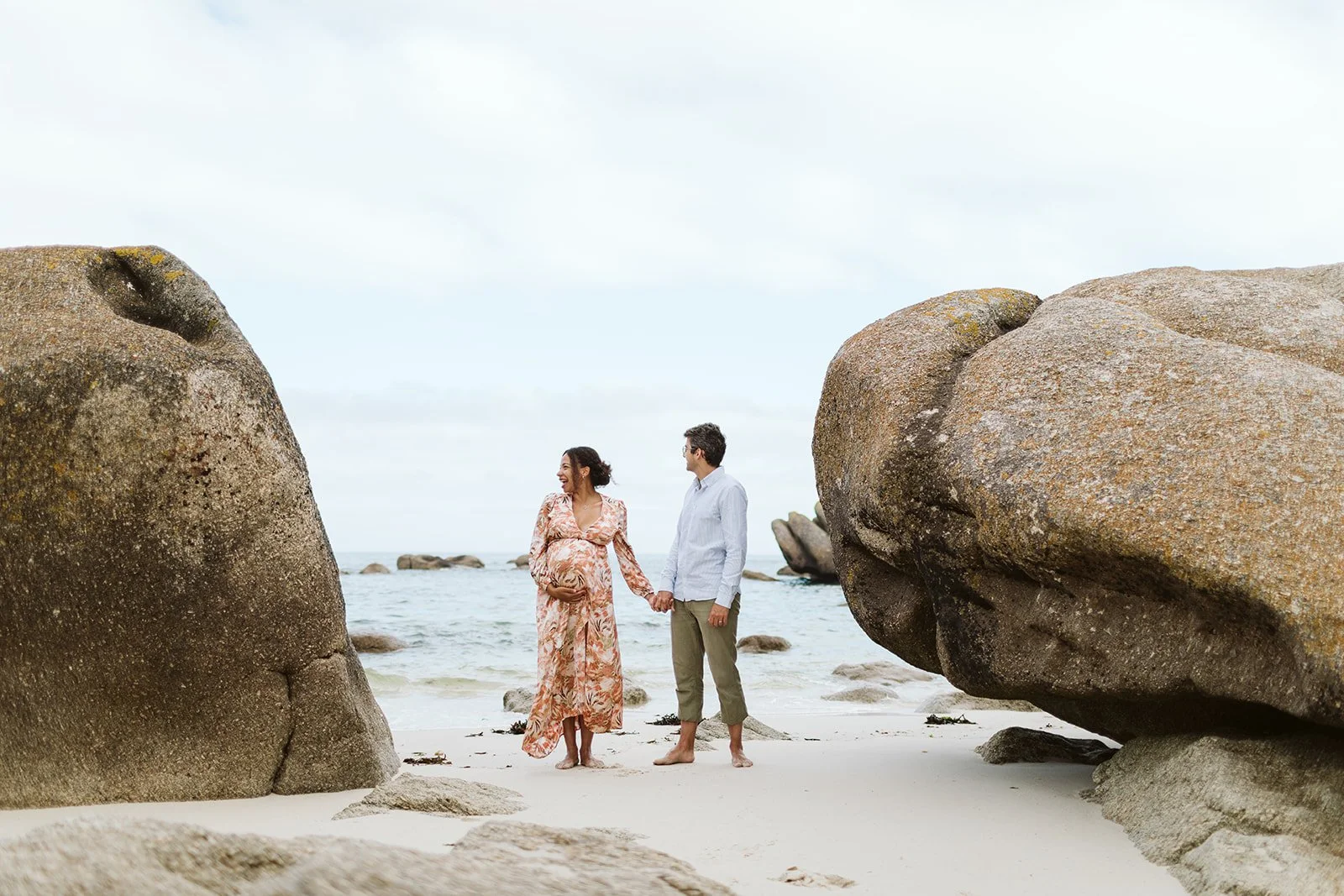 Un couple tenant la main sur une plage avec de gros rochers et la mer en arrière-plan, la femme enceinte portait une robe longue en floral.