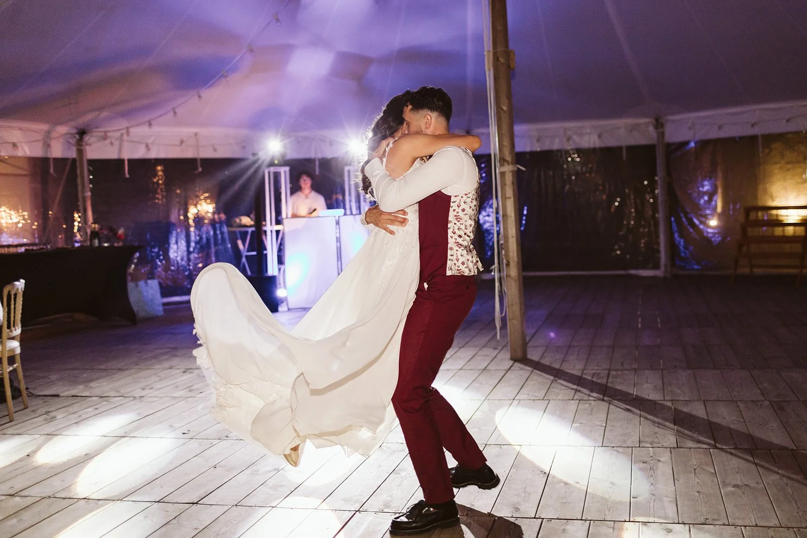 Un couple en habits de mariage danse lors de leur mariage dans une tente. La mariée porte une robe blanche et le marié un costume bordeaux.