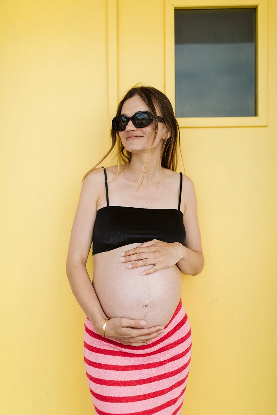 Une femme enceinte portant des lunettes de soleil, un haut noir et une jupe à rayures roses et rouges, qui se tient devant un mur jaune avec une petite fenêtre.