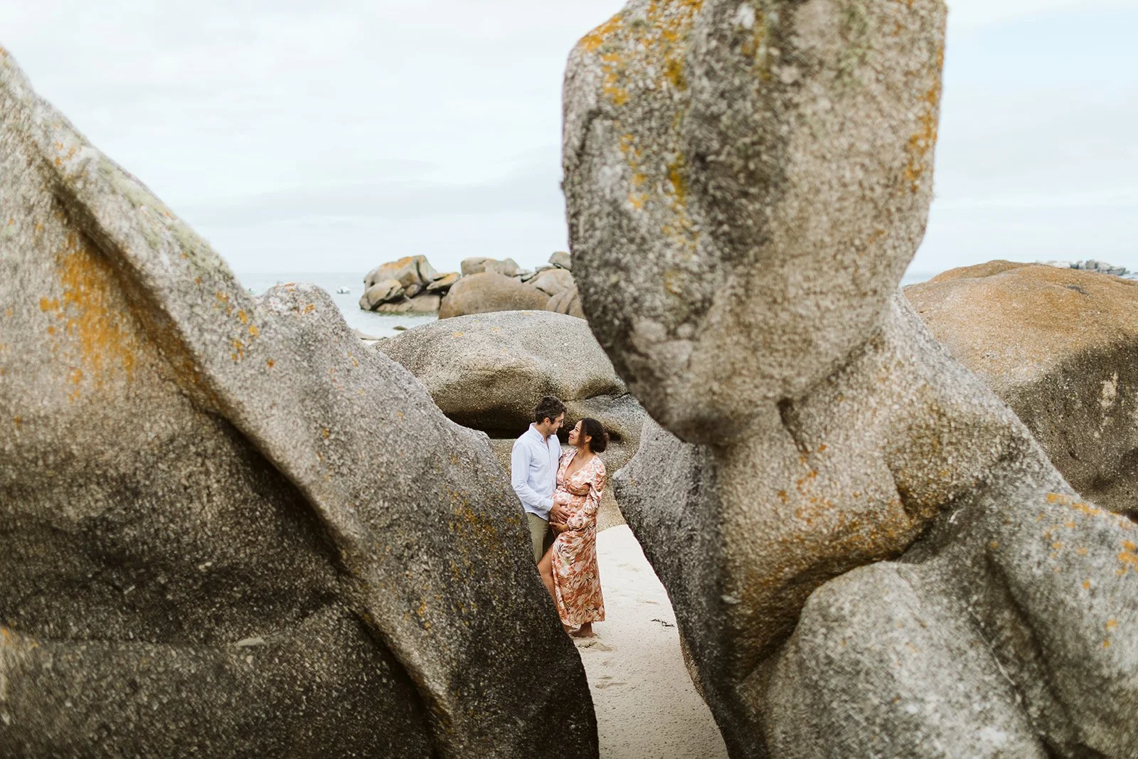 Un couple debout sur une plage, entouré de grosses roches, en train de se regarder tendrement.