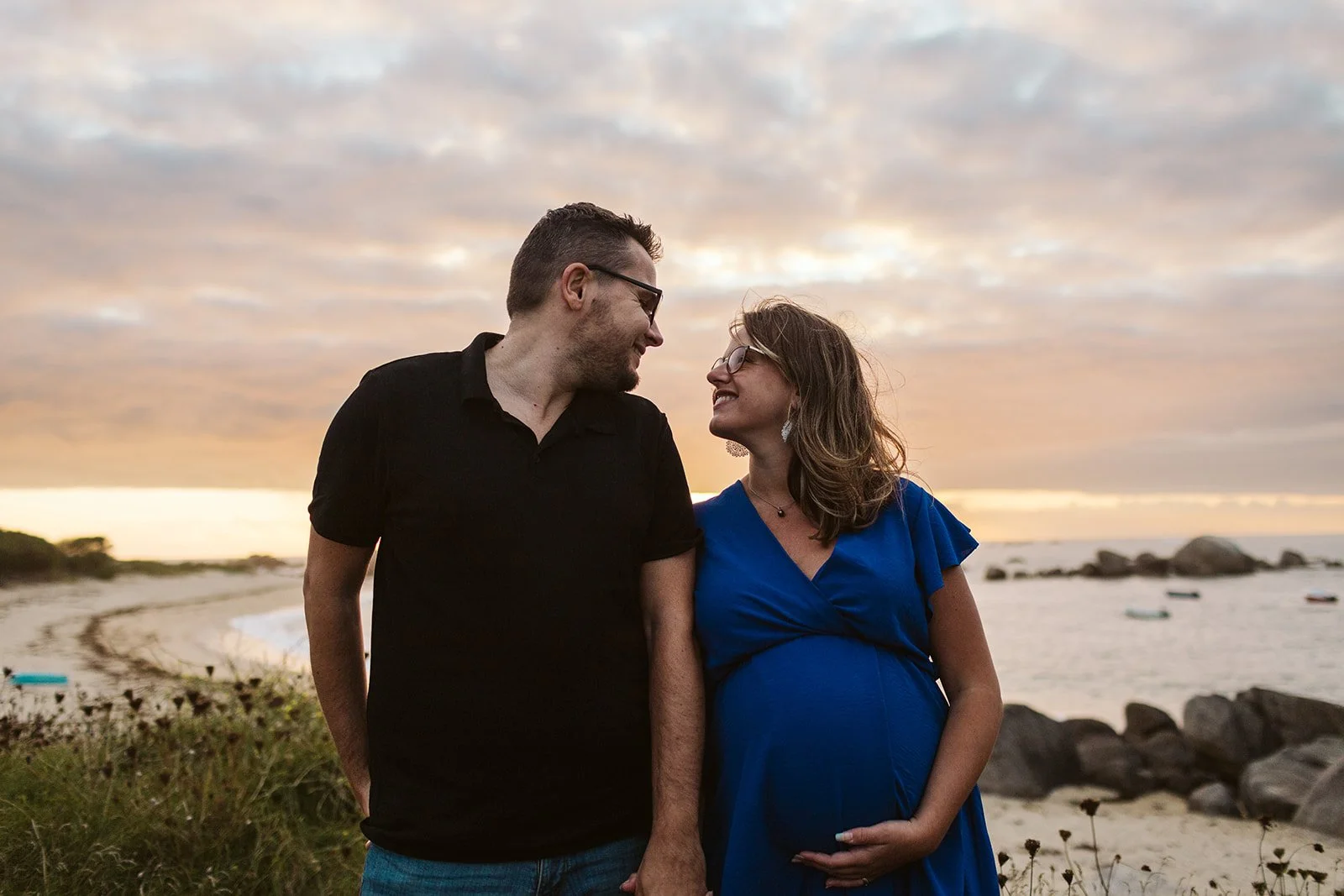Un couple qui regarde et sourit l'un à l'autre sur une plage au coucher du soleil, la femme enceinte porte une robe bleue et tient son ventre.