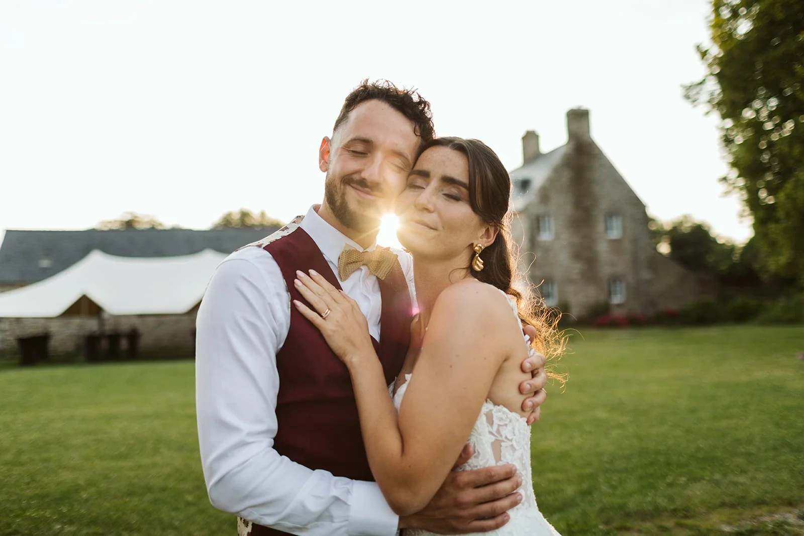 Un couple marié en embrassade lors de leur mariage en extérieur, avec un château en pierre en arrière-plan et le soleil couchant derrière eux.