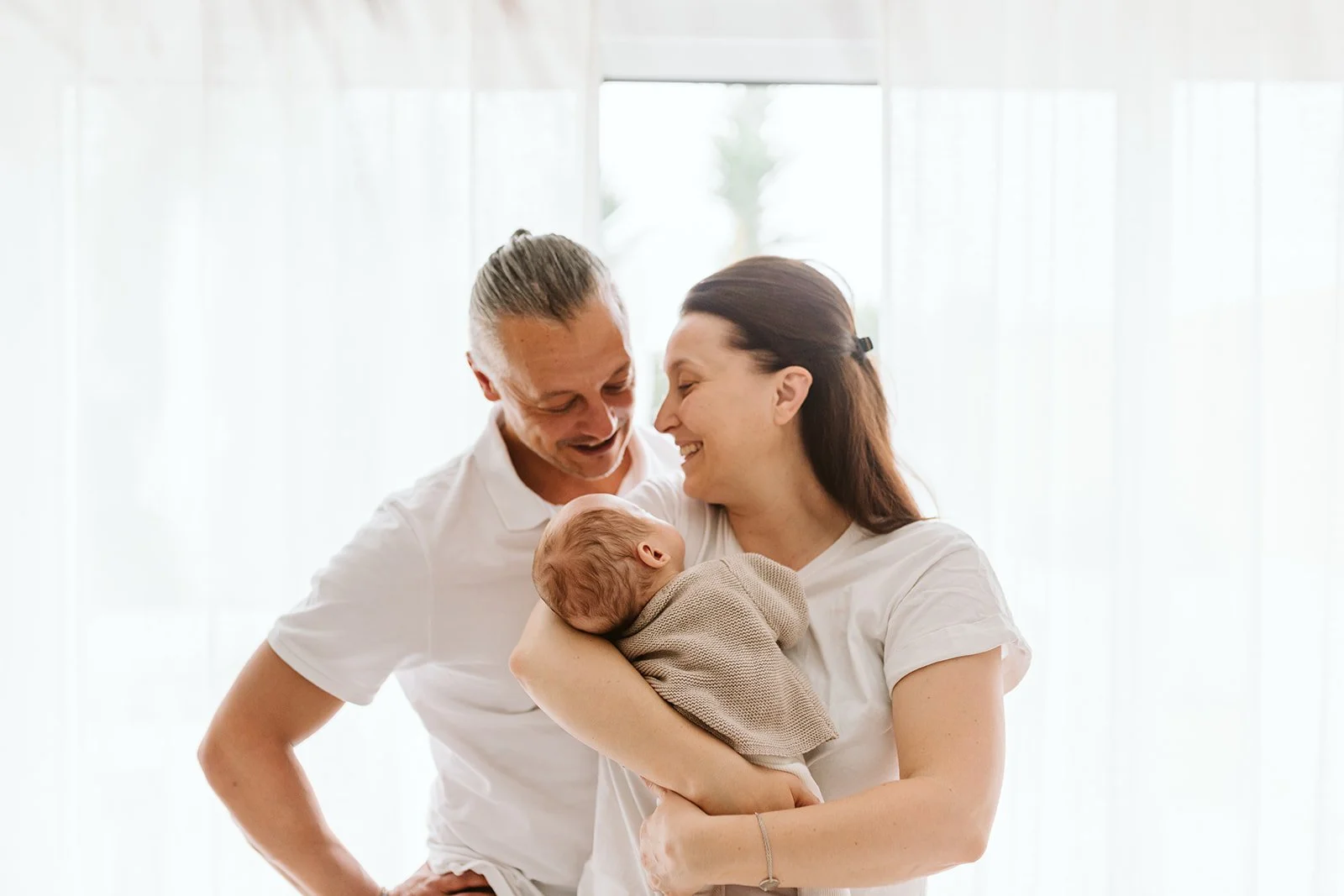Une famille heureuse avec un bébé, la mère et le père sourient devant une fenêtre lumineuse.