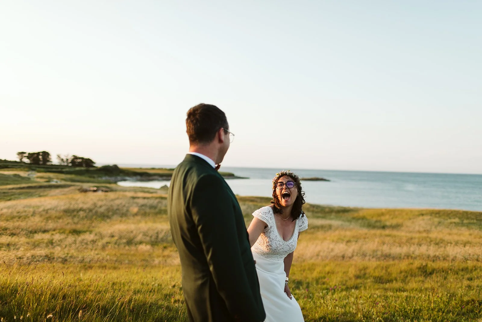 Un couple lors d'un mariage en plein air près de la mer. La femme rit avec enthousiasme, portant une robe blanche et des lunettes. L'homme porte un costume noir avec une chemise blanche. Le paysage est composé d'herbes hautes et de l'océan en arrière