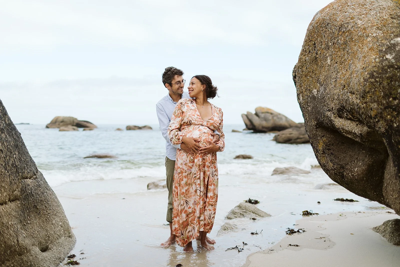 Un couple, dont la femme enceinte, se tient sur la plage entre de grandes roches, avec l'océan en arrière-plan, partageant un moment heureux et complice.