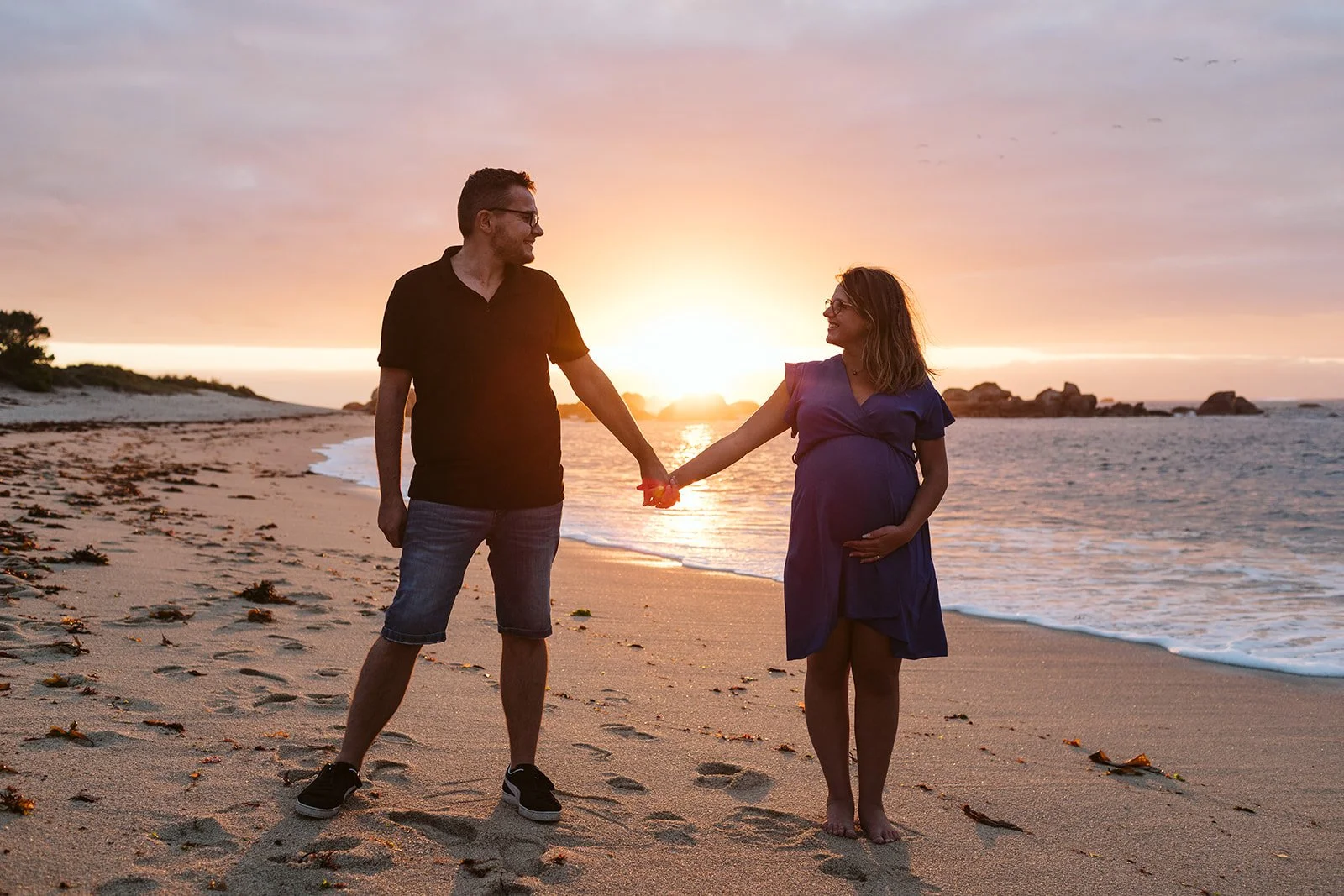 Un couple marchant sur la plage au coucher du soleil, la femme enceinte tenant la main de l'homme.