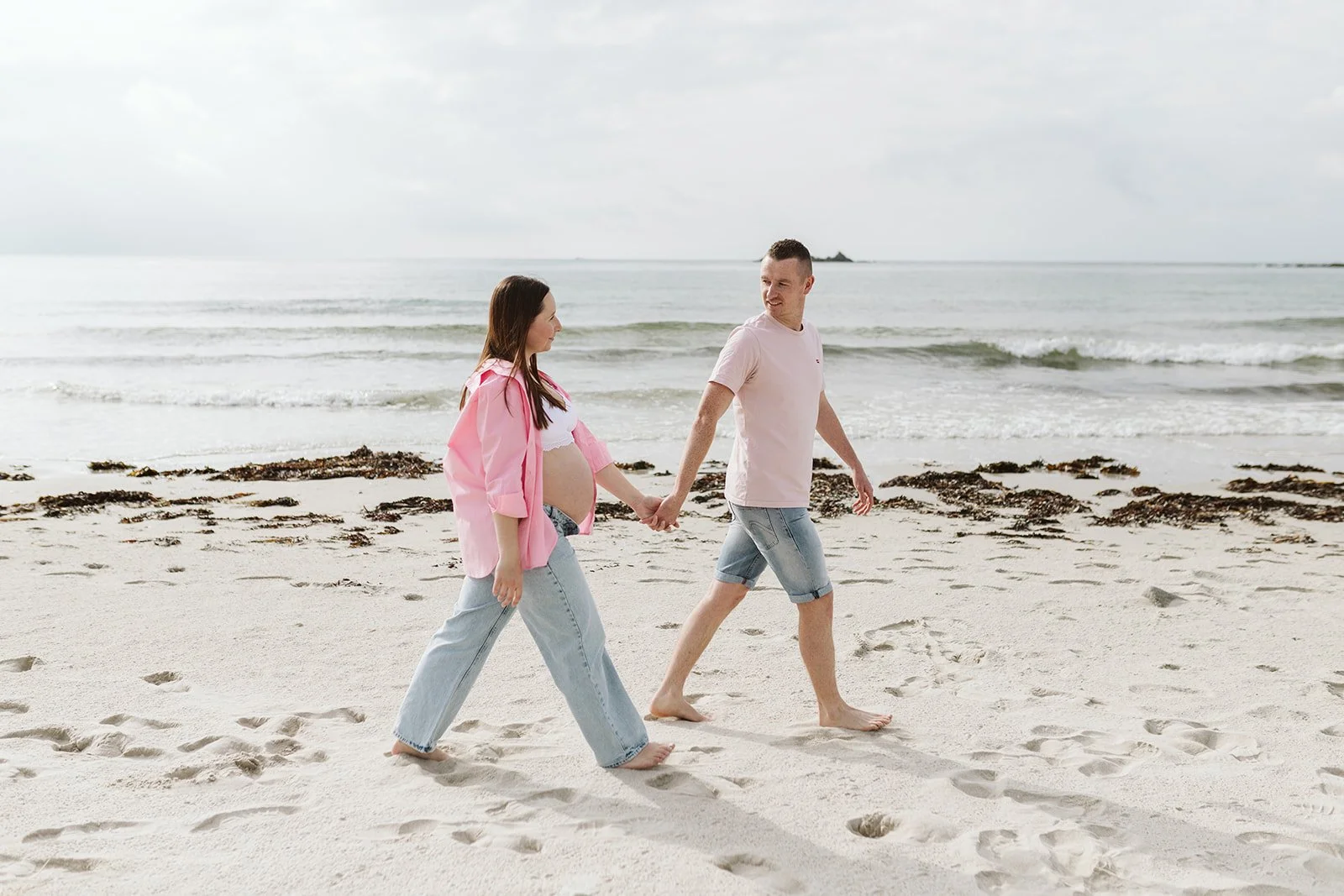 Un couple marche sur la plage, la femme est enceinte, ils se tiennent la main, face à la mer.