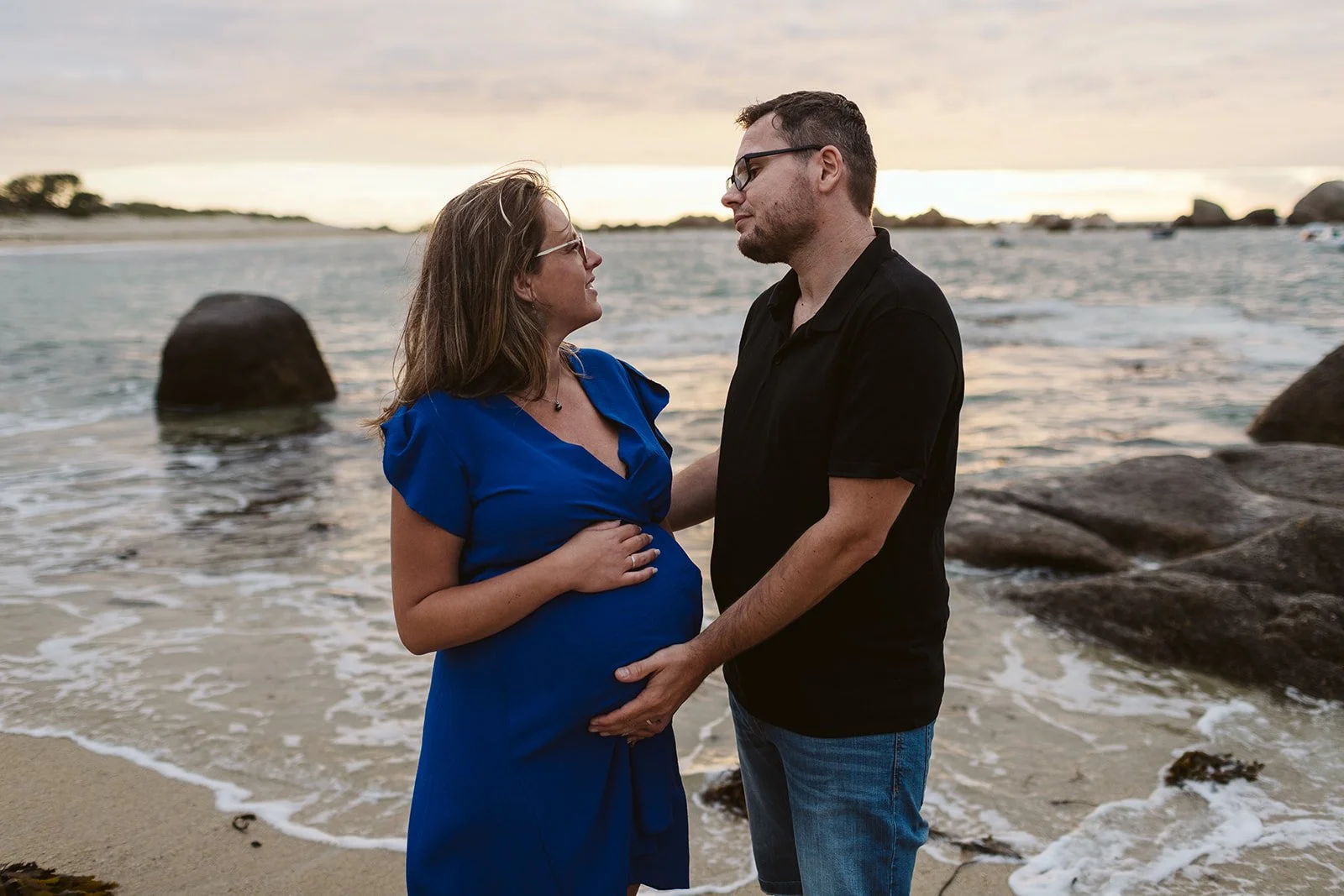 Une femme enceinte en robe bleue et un homme en chemise noire face à face sur la plage au coucher du soleil, tenant doucement son ventre tout en se regardant dans les yeux.