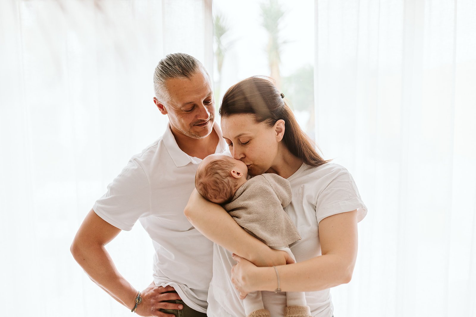 Une famille de trois personnes, un homme, une femme et un bébé, dans une pièce lumineuse. La femme tient le bébé dans ses bras et l'embrasse sur le front, tandis que l'homme regarde avec tendresse.