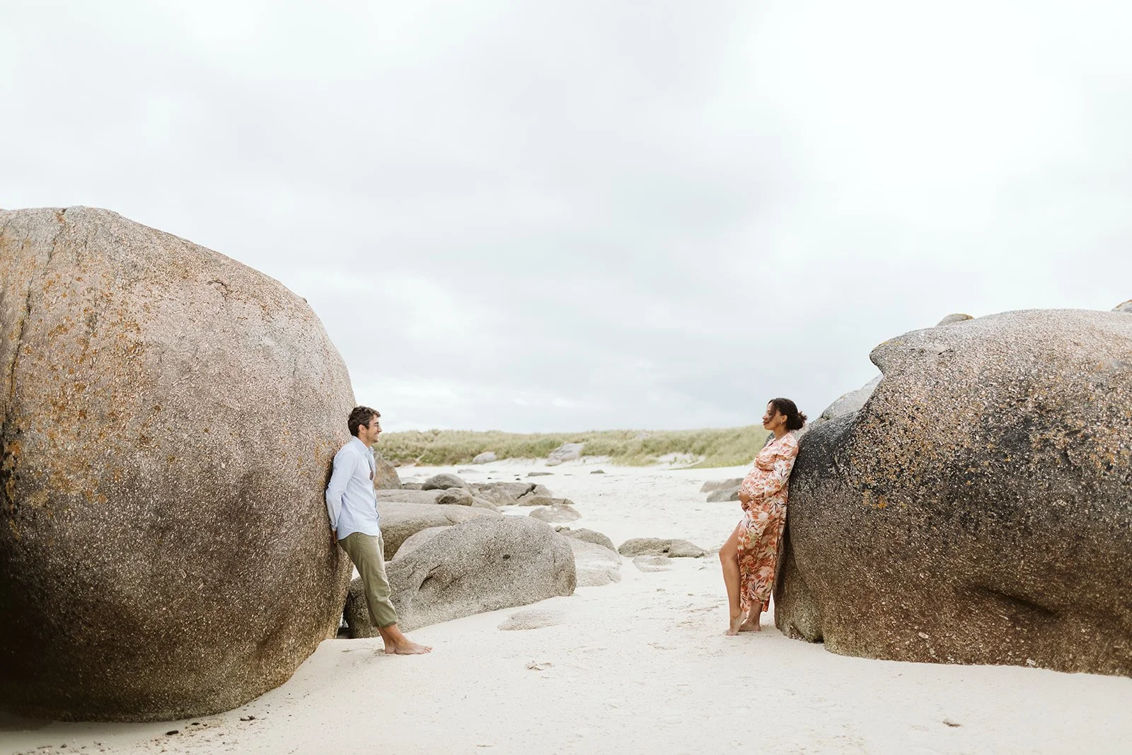 Un couple regarde l'un l'autre entre deux gros rochers sur une plage, avec un ciel nuageux en arrière-plan.
