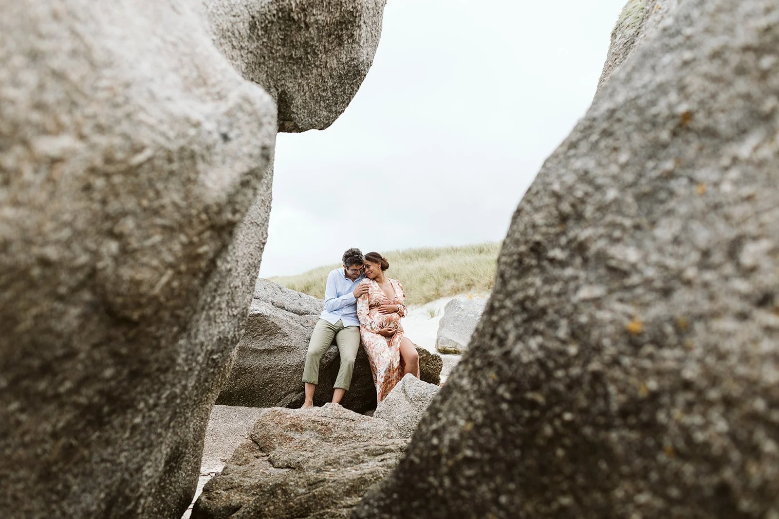 Un couple assis sur des rochers à la plage, entouré de gros rochers, avec de l'herbe en arrière-plan, moment intime et romantique