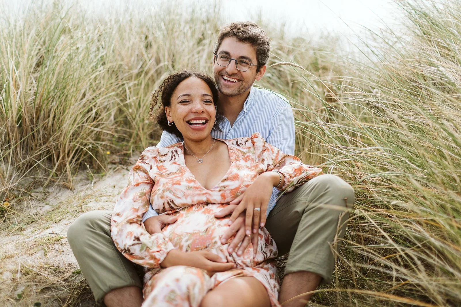 Un couple souriant, assis dans un pré de hautes herbes, dont la femme est enceinte, lors d'une séance photo en extérieur.