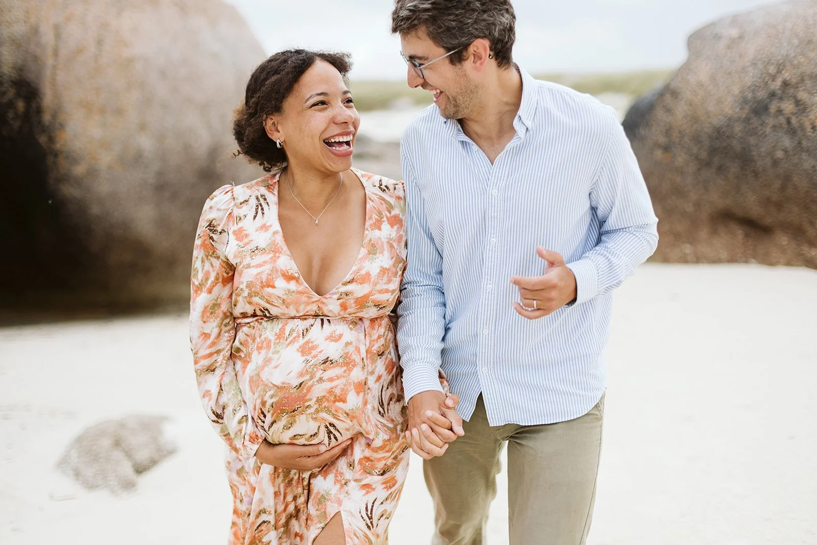 Un couple heureuse se tenant la main et marchant sur la plage, il y a de gros rochers en arrière-plan.
