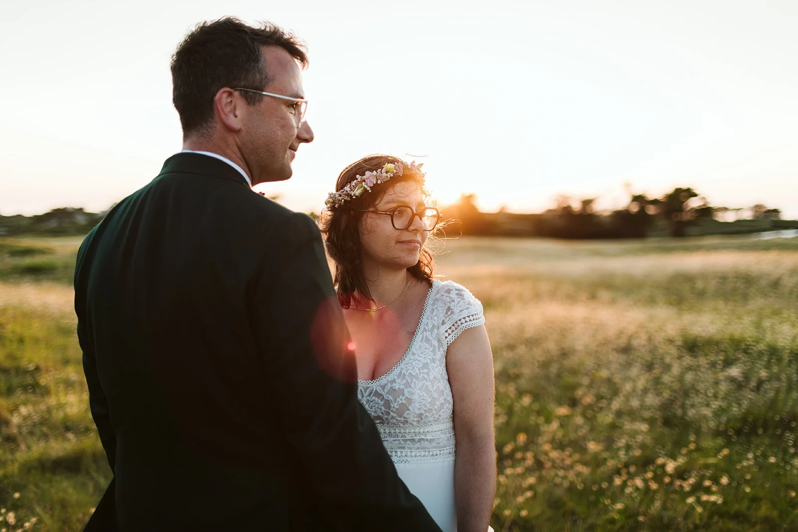 Un couple lors d'un mariage en plein air au coucher du soleil, la femme porte une couronne de fleurs et des lunettes, l'homme porte un costume noir.