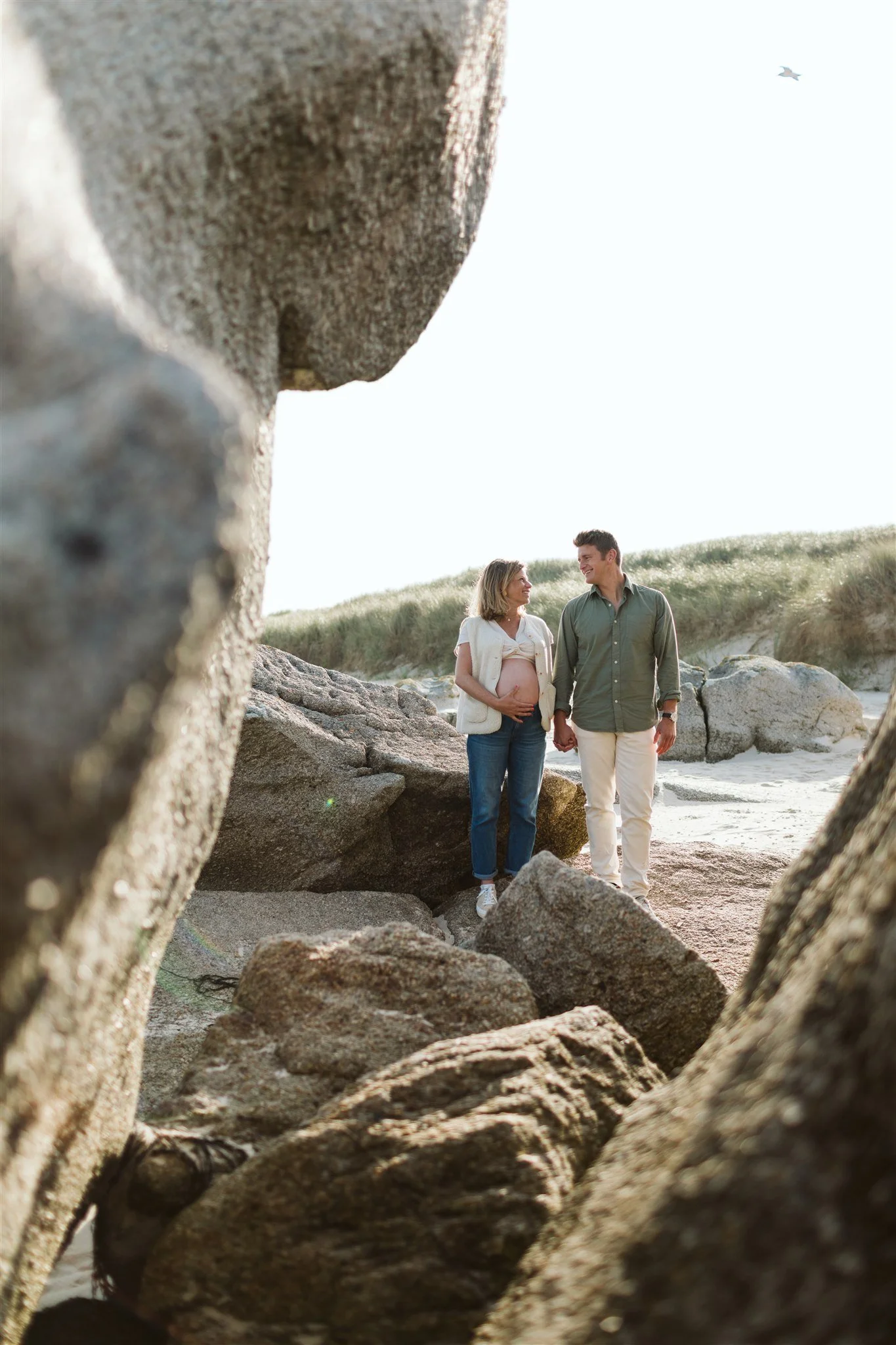 Un couple marche sur la plage, l'homme tient la main de la femme qui est enceinte. Ils se regardent souriant. La photo est prise à travers des rochers, avec un ciel clair en arrière-plan.