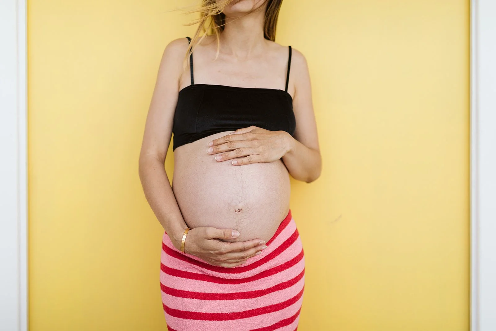 Femme enceinte portant un top noir et une jupe à rayures roses et rouges, tenant son ventre contre un mur jaune.